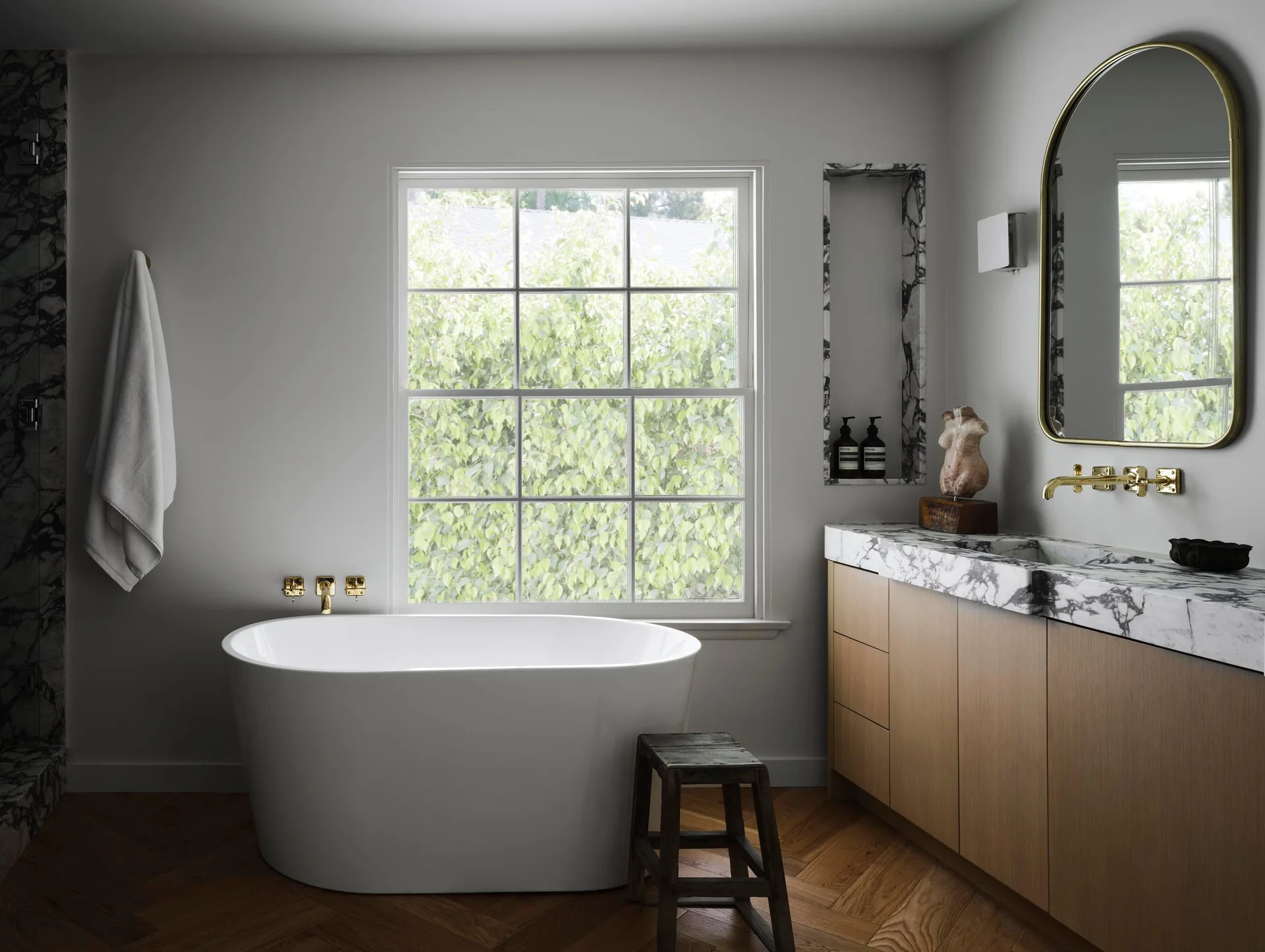 A modern bathroom features a white freestanding bathtub by a large window, a wood vanity with a thick marble countertop, and herringbone wood floors.