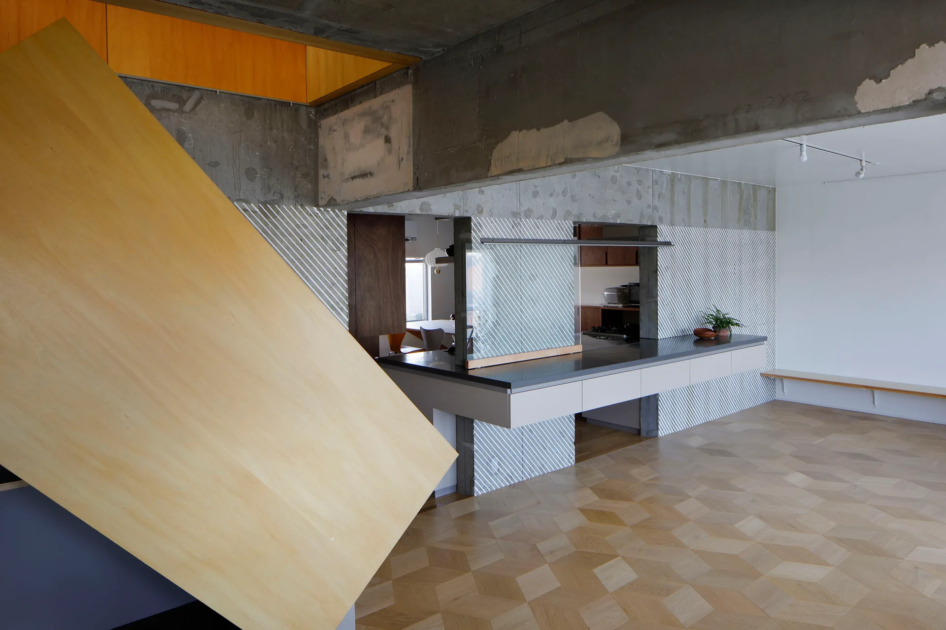 An open-concept kitchen features a long gray counter, exposed raw concrete ceiling beams, diagonal striped walls, and geometric parquet flooring.