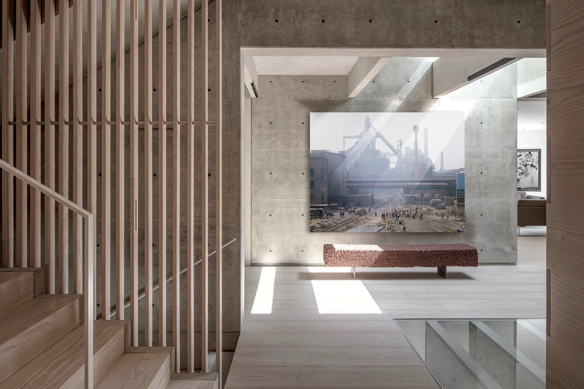 A minimalist interior features exposed concrete walls, a light wood staircase behind a vertical slat screen, and a textured red-brown bench under wall art.