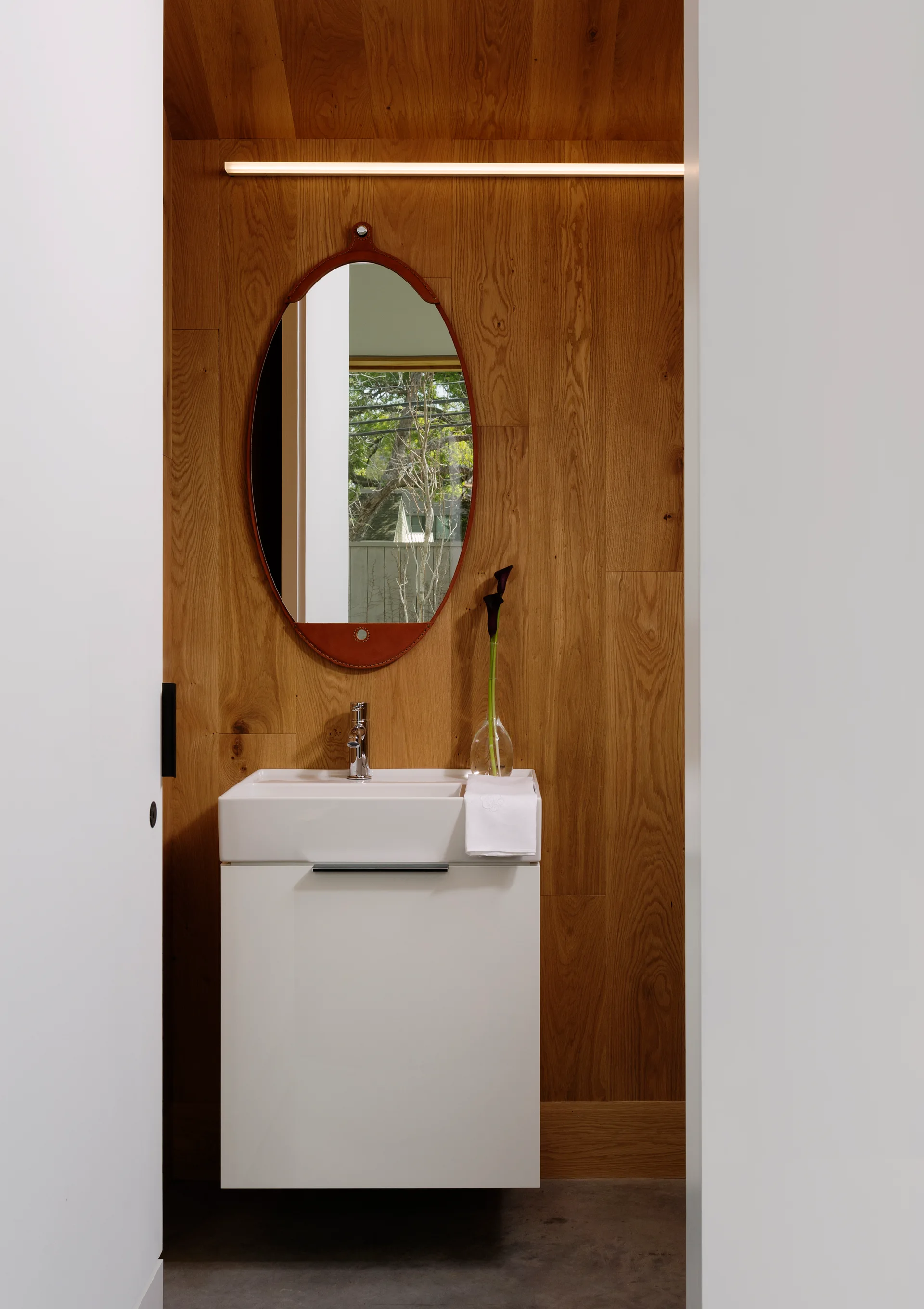 A minimalist powder room features a white floating vanity against a vertical wood paneled wall with an oval leather trimmed mirror.