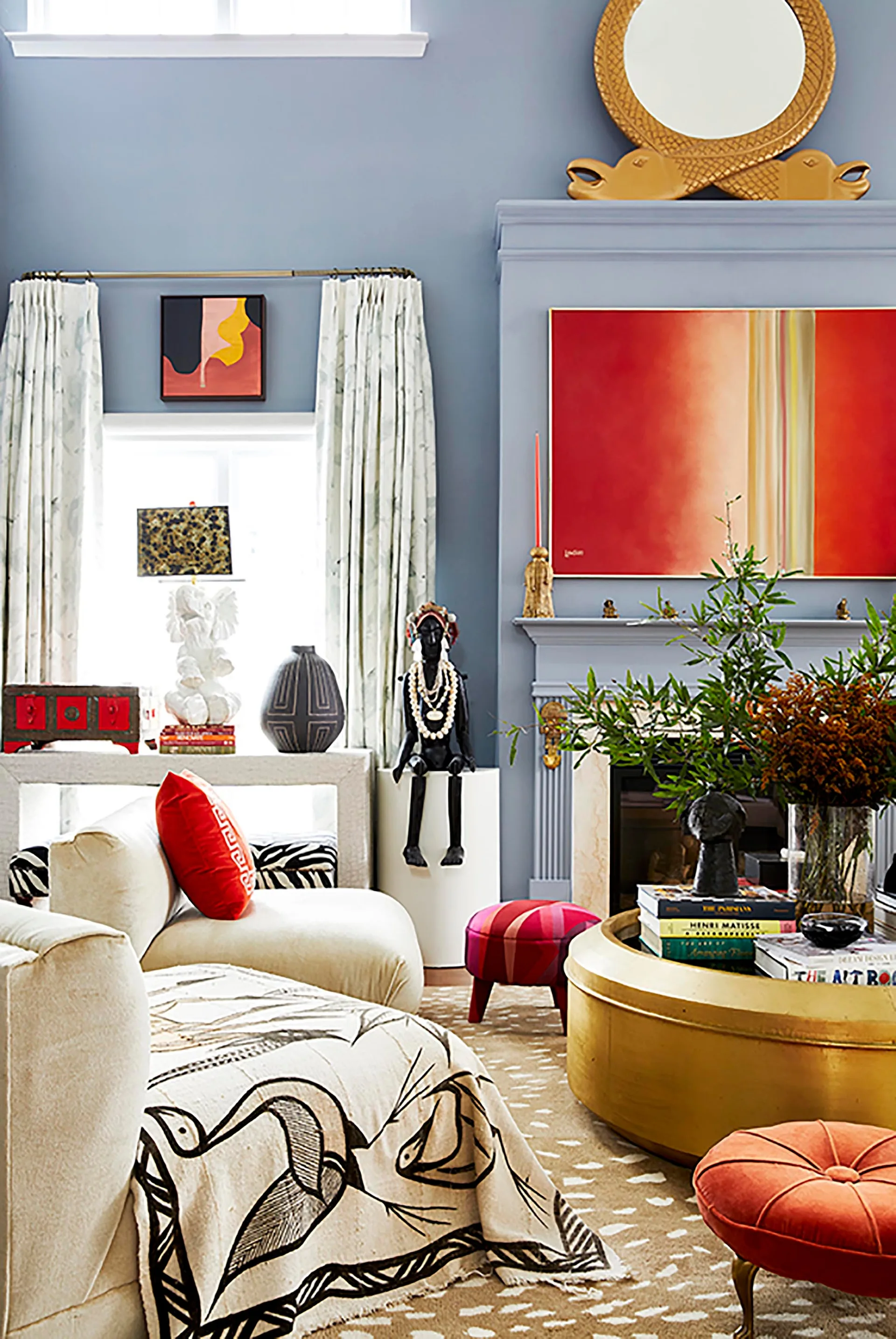 A blue living room features a painted blue fireplace mantel, a gold cylindrical coffee table, a red abstract painting, and patterned furniture.