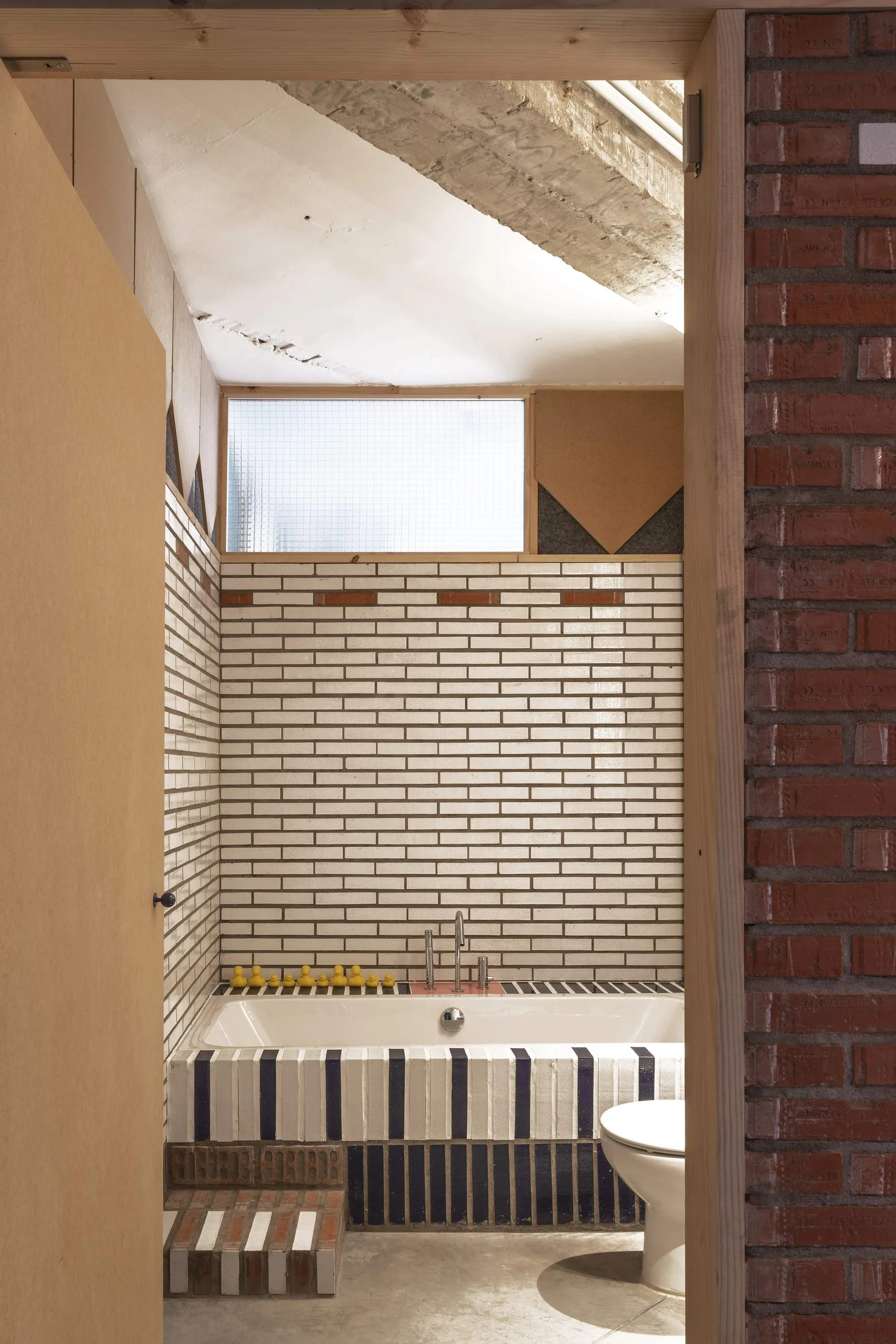 A bathroom view featuring a white bathtub clad in navy and white striped tile, surrounded by white brick walls and a raw brick entryway.