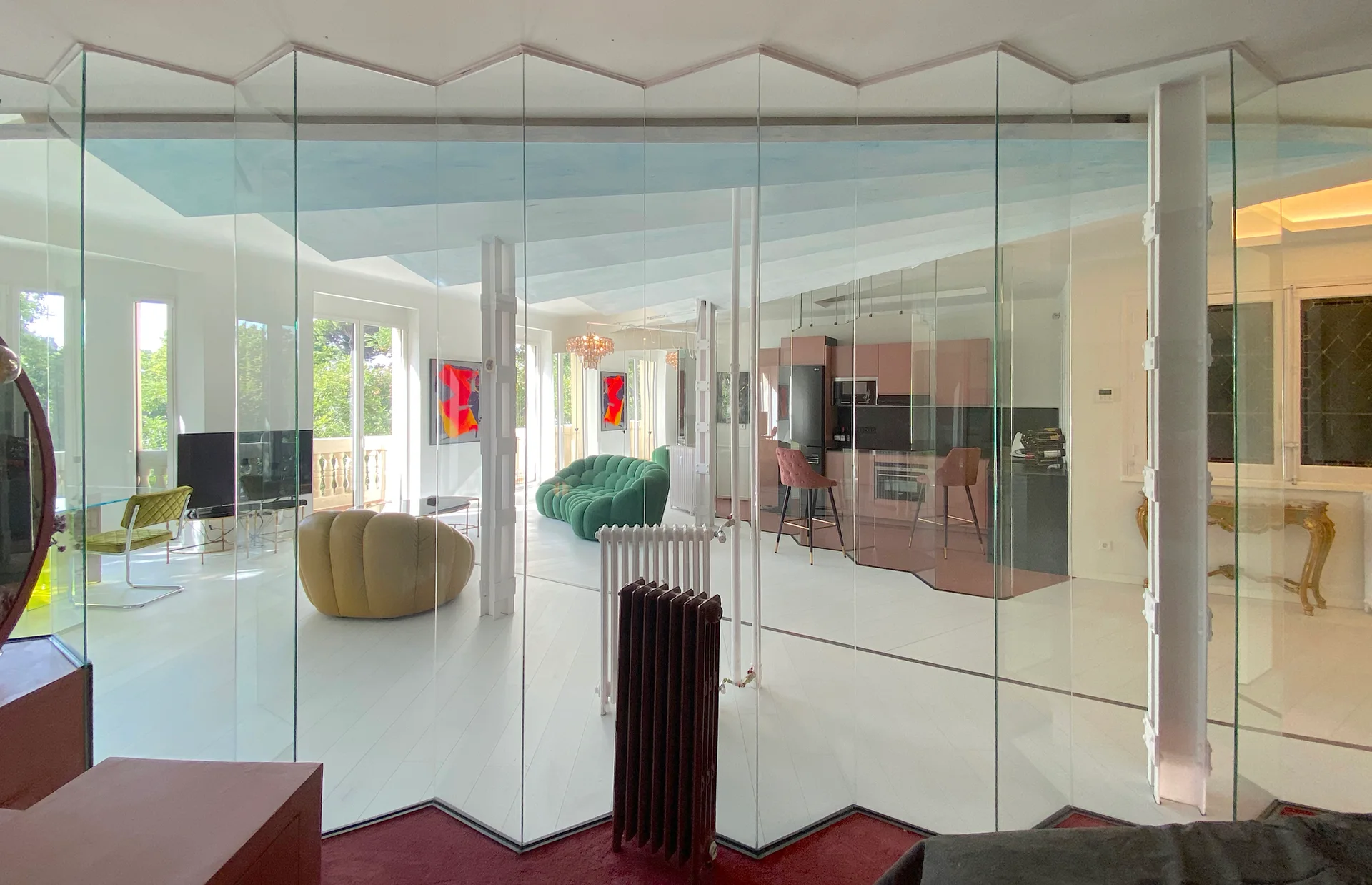 An open-plan living room with a distinctive zigzag glass partition, a green velvet sofa, white floors, and a pink kitchen in the background.