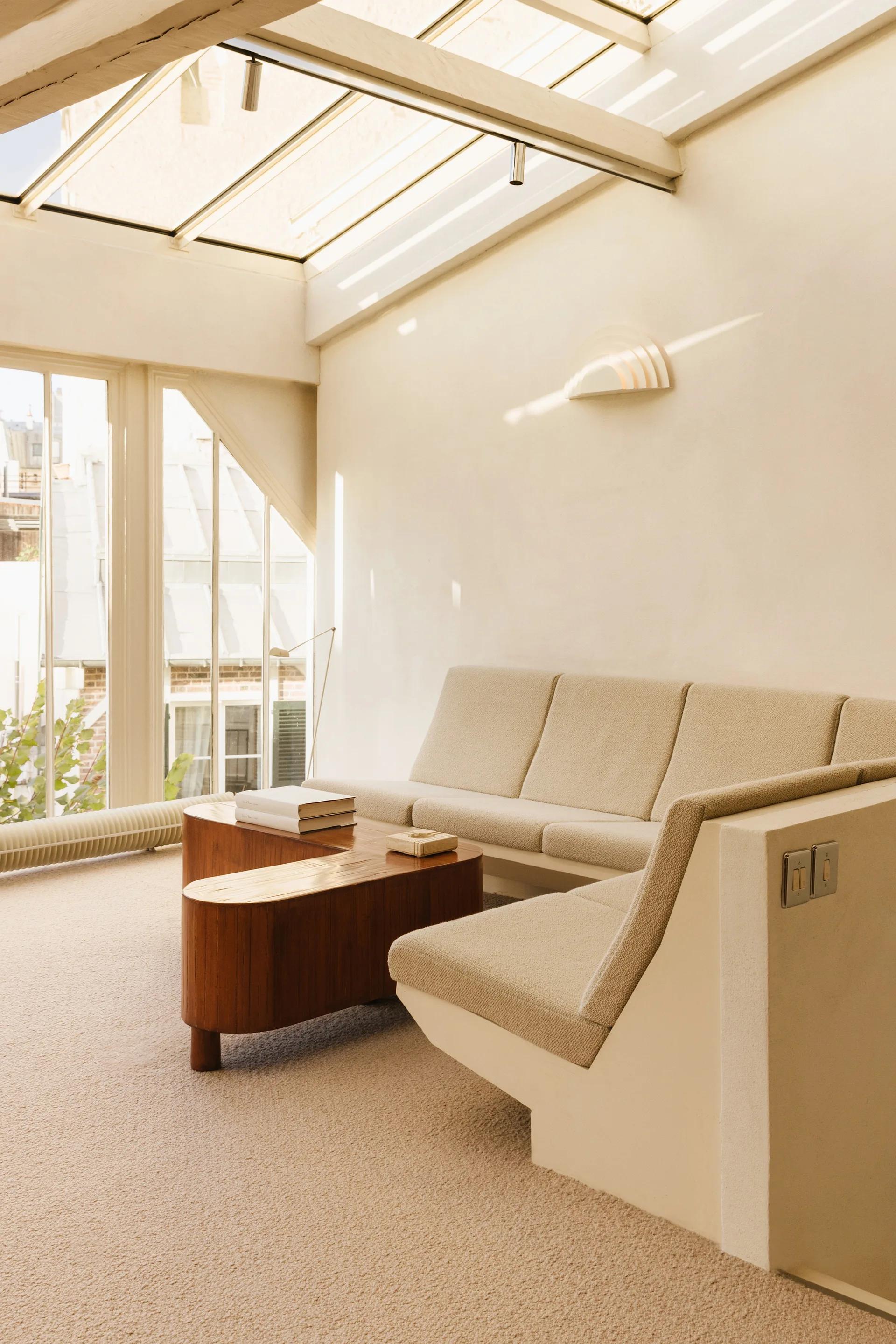 A minimalist living room features a beige built-in sofa, a curved wood coffee table, and a large angled glass skylight overhead.