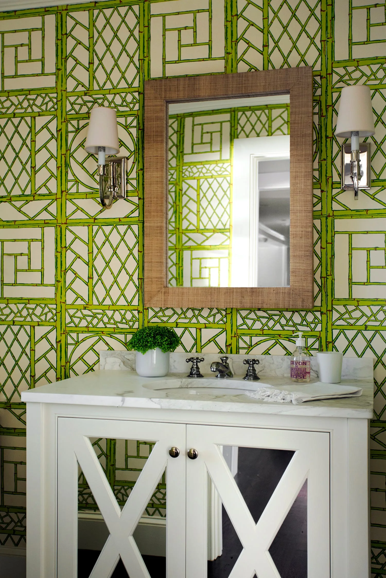 A white bathroom vanity with a marble top stands against green bamboo lattice wallpaper, below a mirror with a woven frame.