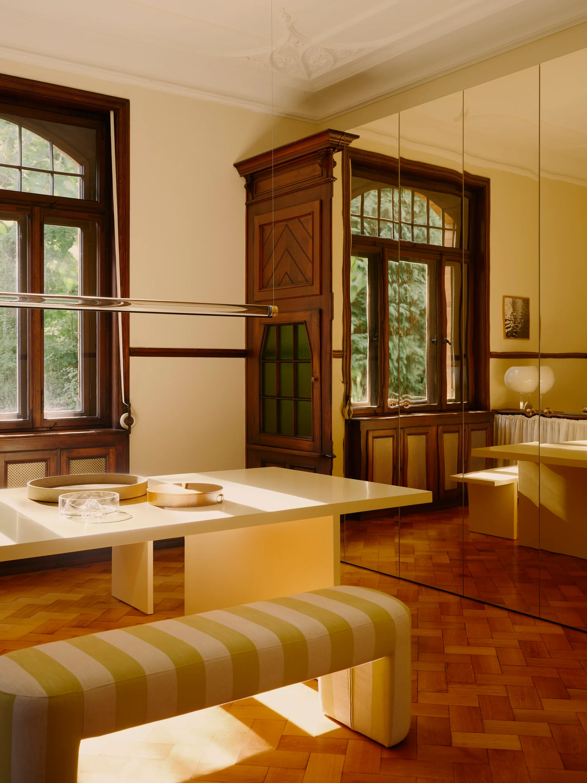 A modern cream dining table and a striped bench sit in a room with a large mirrored wall, herringbone wood floors, and dark wood-framed windows.