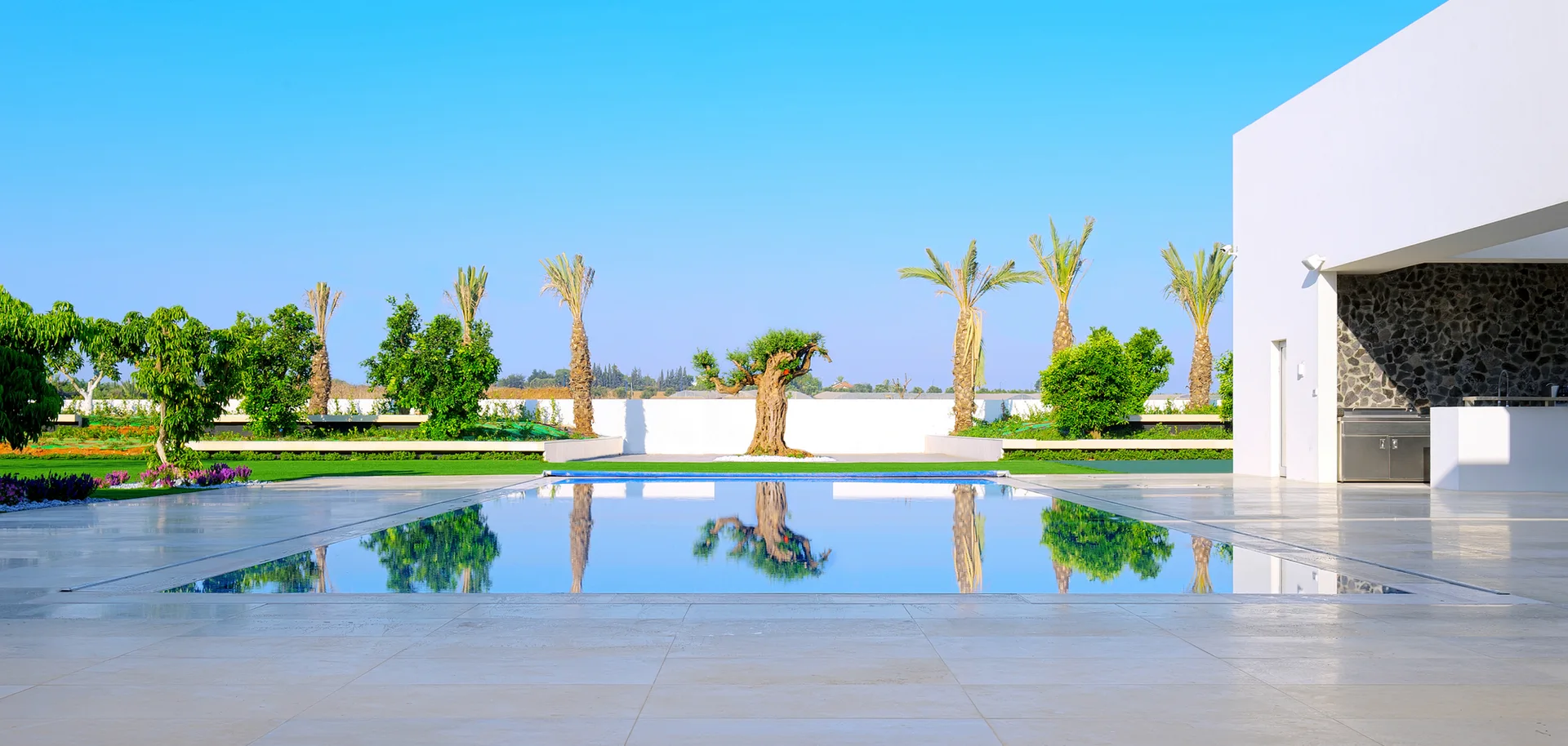 A large rectangular swimming pool reflects the clear blue sky and palm trees on a spacious, light-tiled modern patio next to a white building.