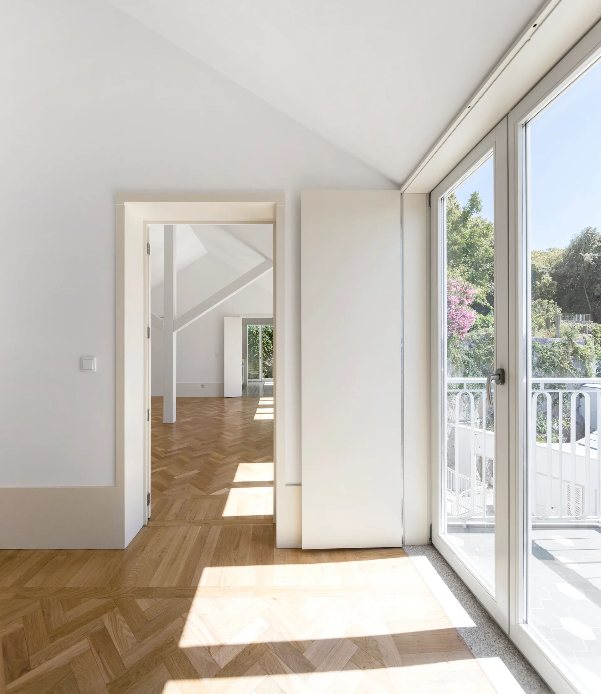 A minimalist white room features herringbone pattern wood flooring, a deep doorway, and a glass balcony door.