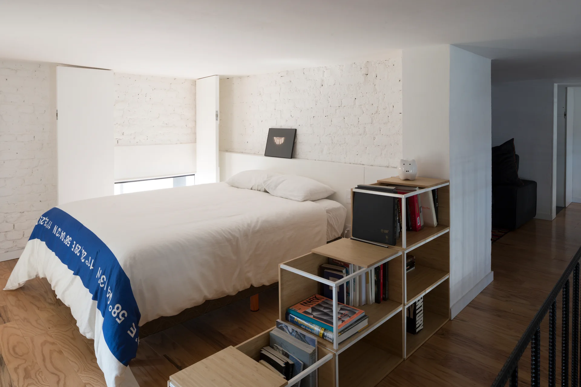 A minimalist loft bedroom features a bed with white bedding, a white painted brick wall, and a low wooden bookshelf acting as a room divider.
