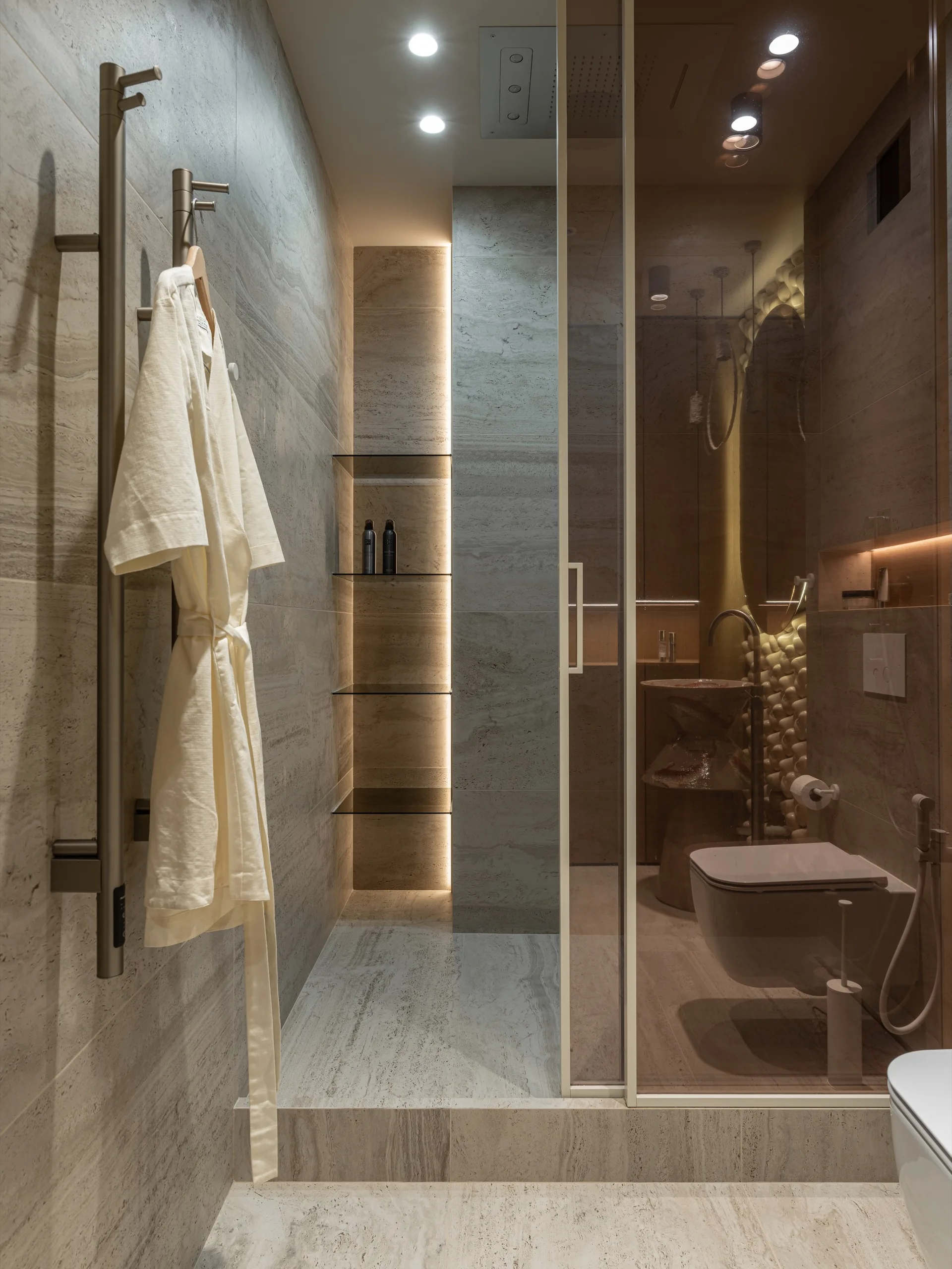A modern bathroom featuring gray stone tiles, a vertical backlit recessed wall niche with glass shelves, a wall-mounted toilet, and a glass partition.