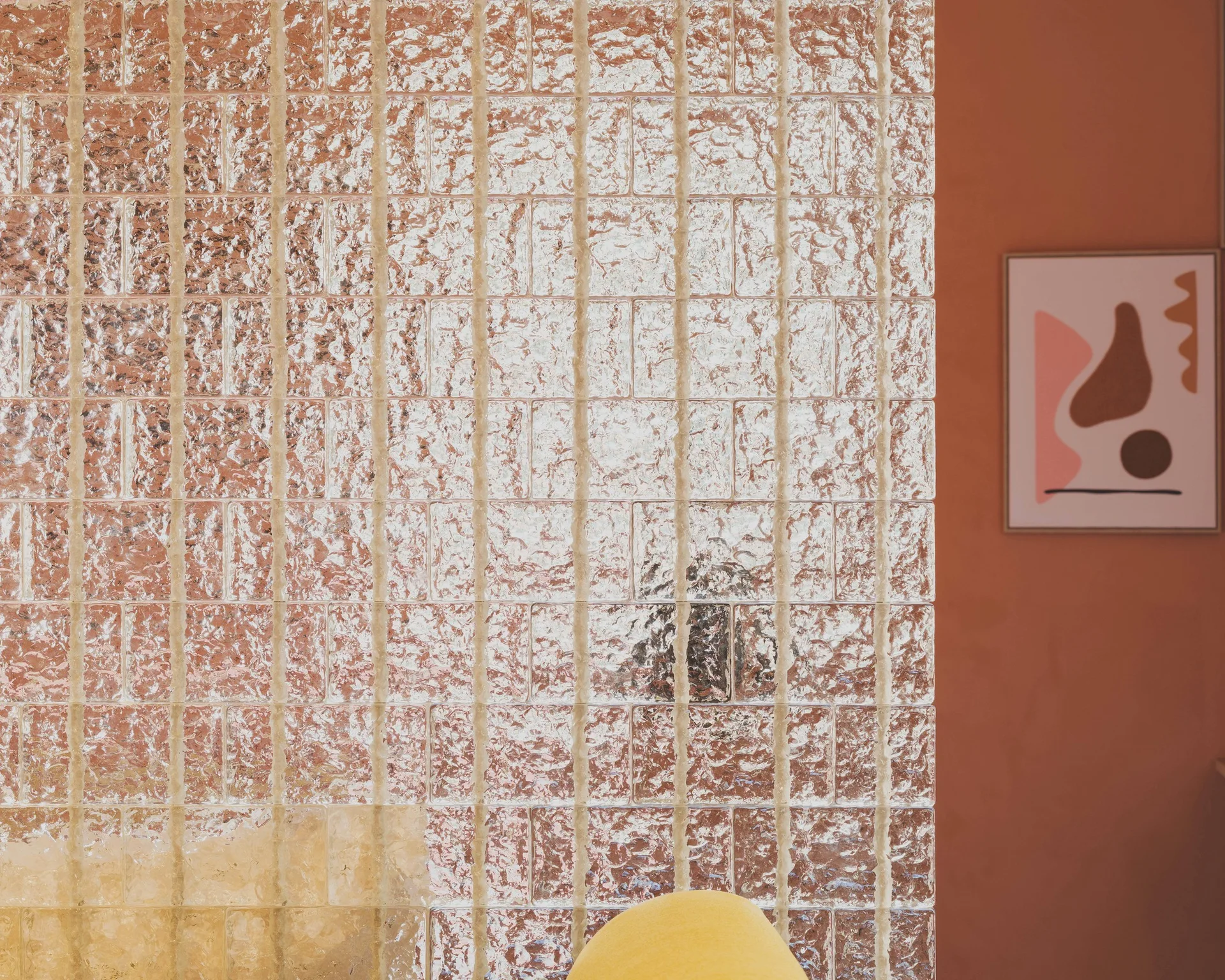A close-up shot of a textured glass block partition wall in front of a warm terra-cotta wall decorated with abstract framed art.