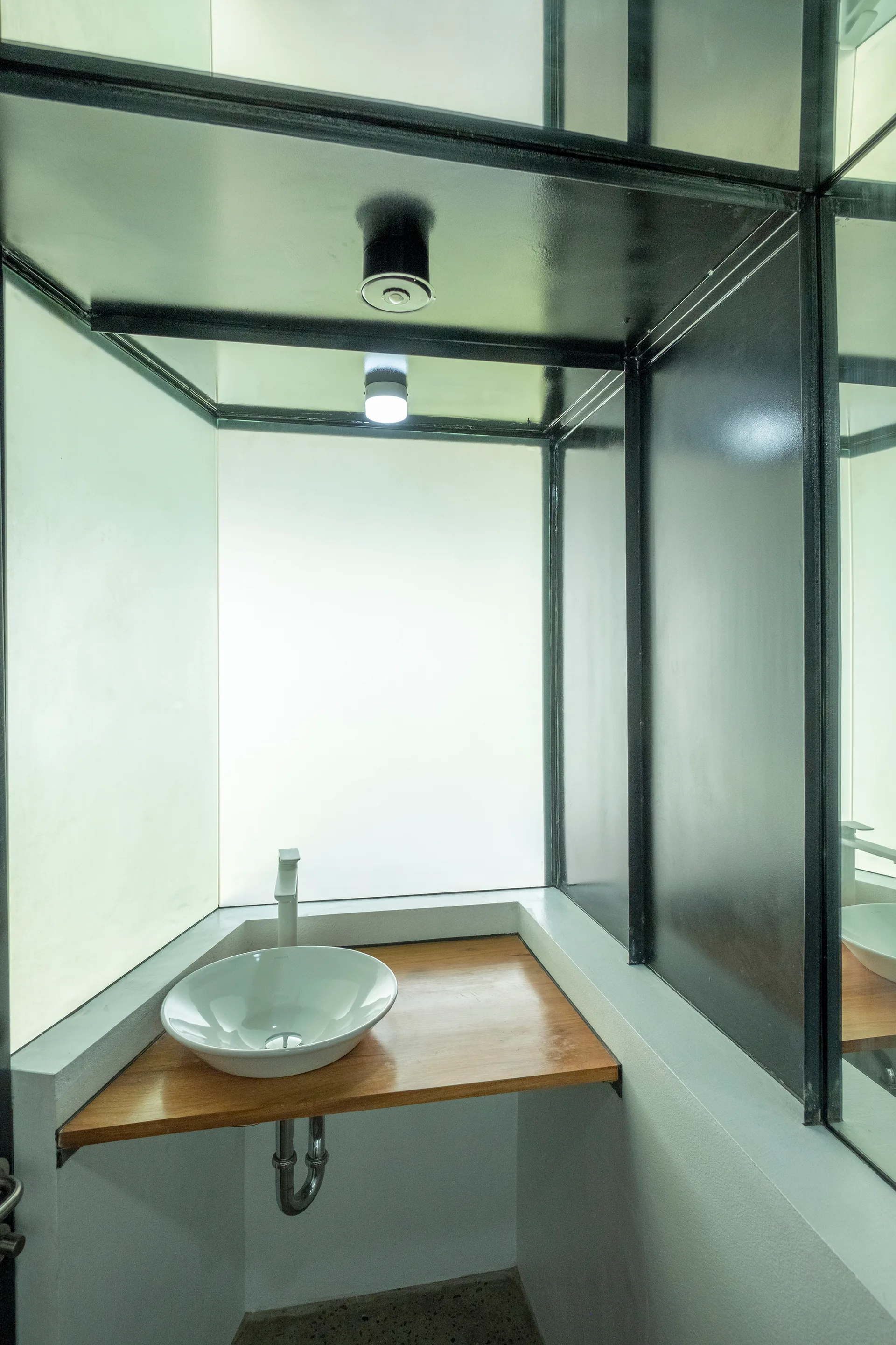 A modern bathroom corner features a white vessel sink on a triangular wooden vanity shelf, surrounded by black-framed frosted glass and mirror panels.