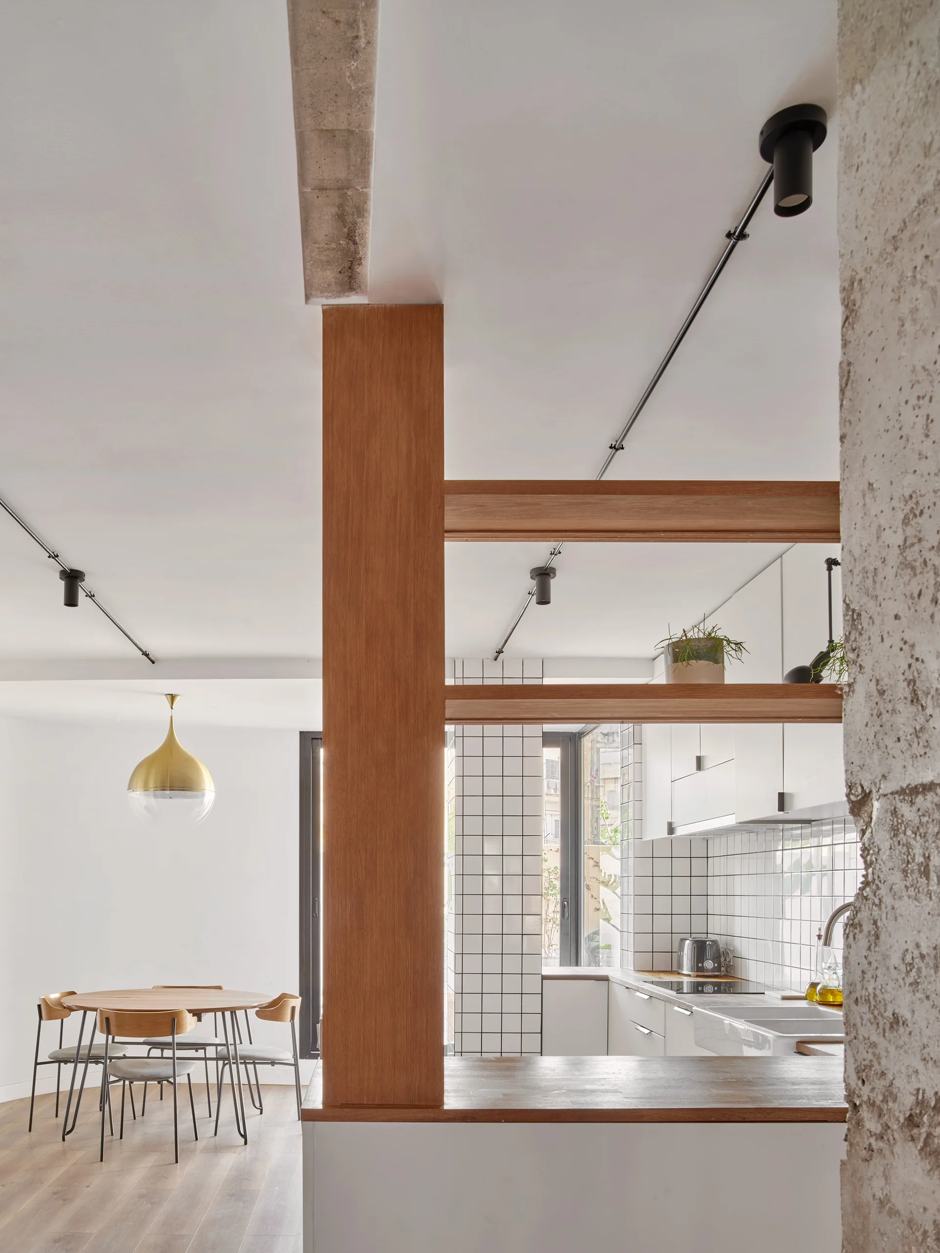 A modern kitchen and dining area features a wood timber divider, white square tile backsplash, and a round wooden dining table under a pendant light.