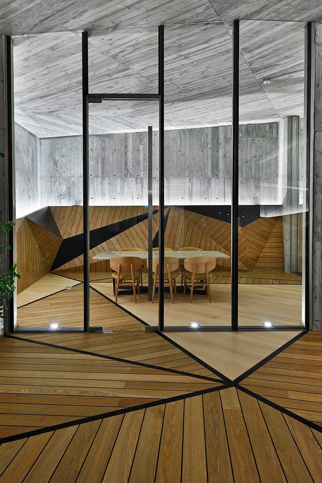 A dining room seen through a glass partition, featuring a wooden table and chairs, geometric-patterned wood flooring, and exposed concrete walls.