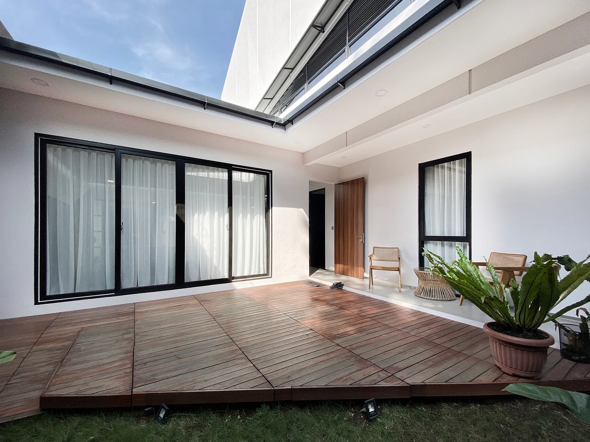 A modern outdoor patio features a wooden deck, white stucco walls, black-framed sliding glass doors, and rattan chairs.