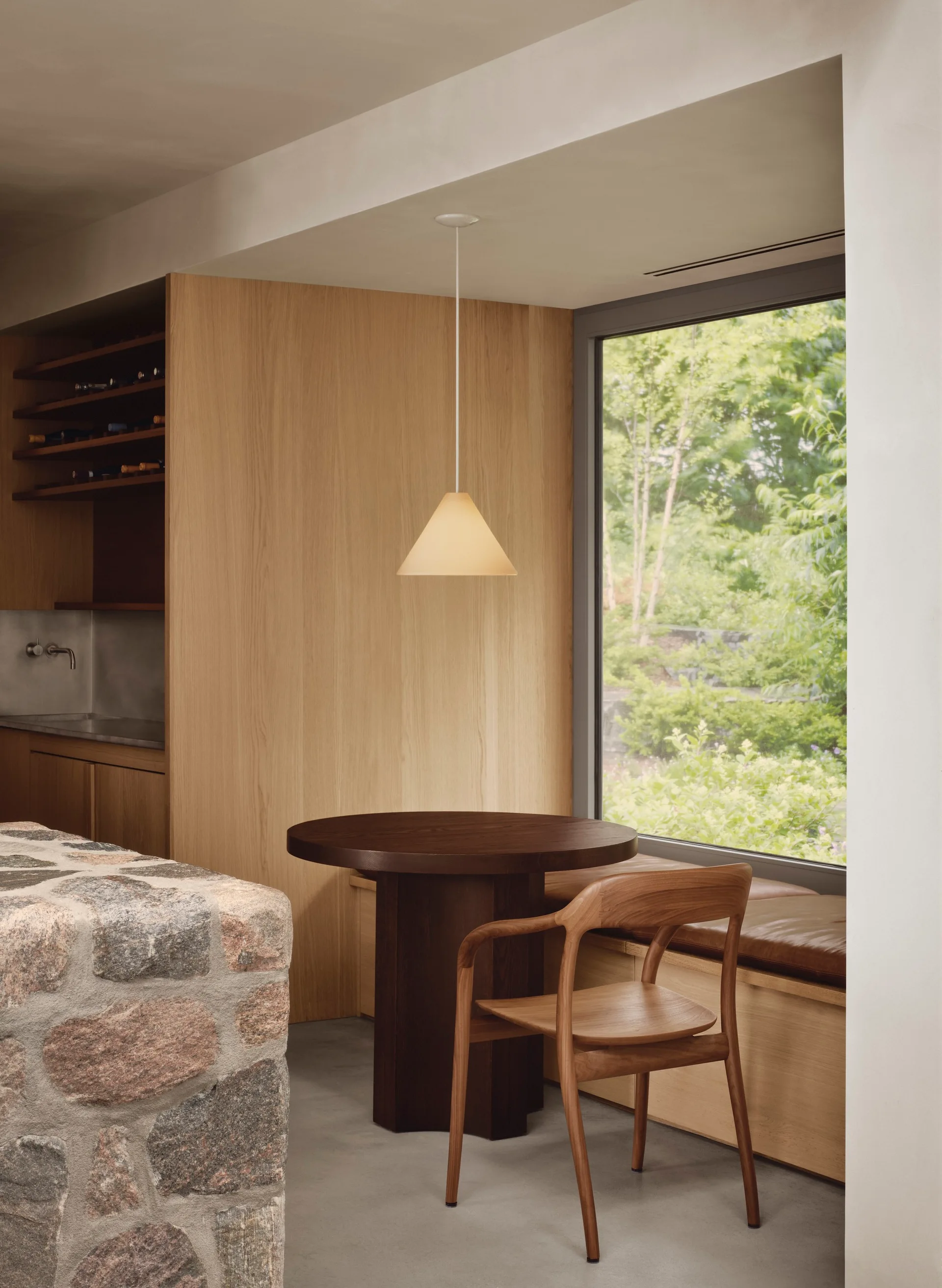 A minimalist dining nook features a dark wood pedestal table, a wooden chair, a built-in window bench, and a textured stone wall in the foreground.