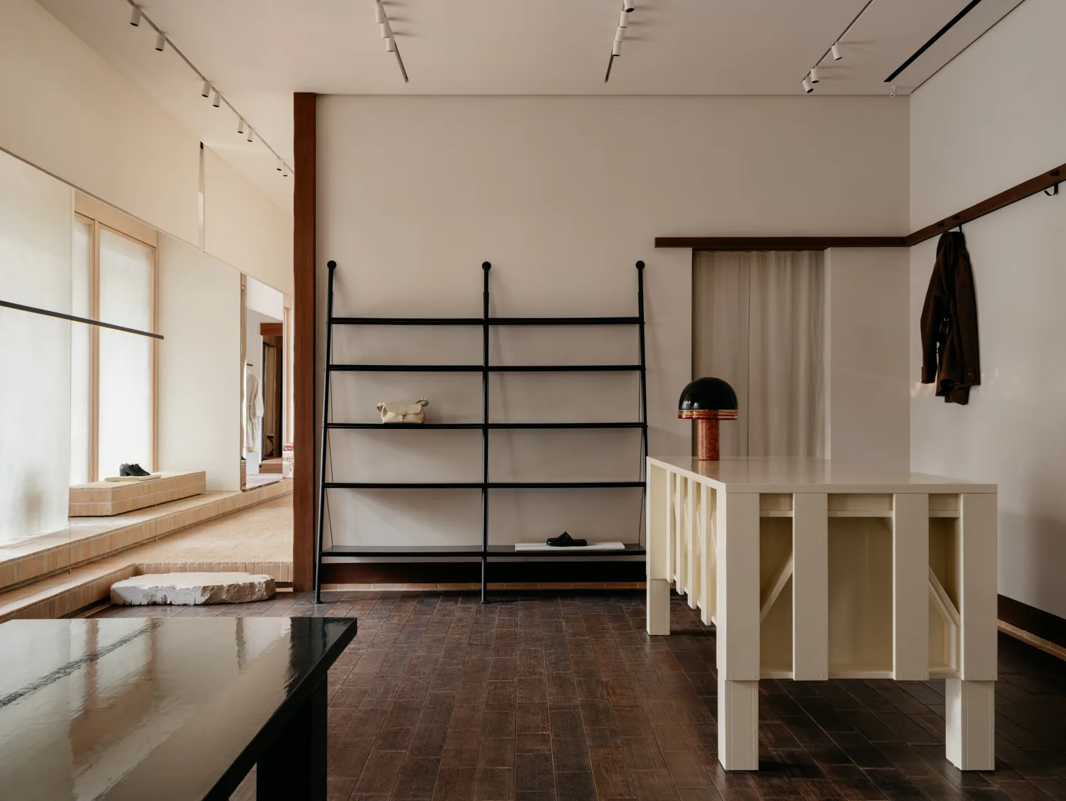 A minimalist showroom interior features dark wood plank flooring, a large cream display table, and a lean-to black metal shelving unit against a plain wall.