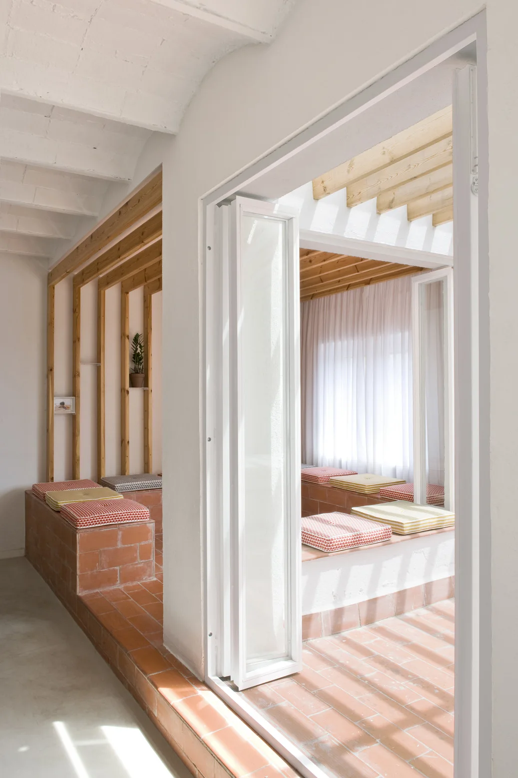 An interior room features built-in red brick benches with patterned cushions on terracotta tile floors near an open white folding door.