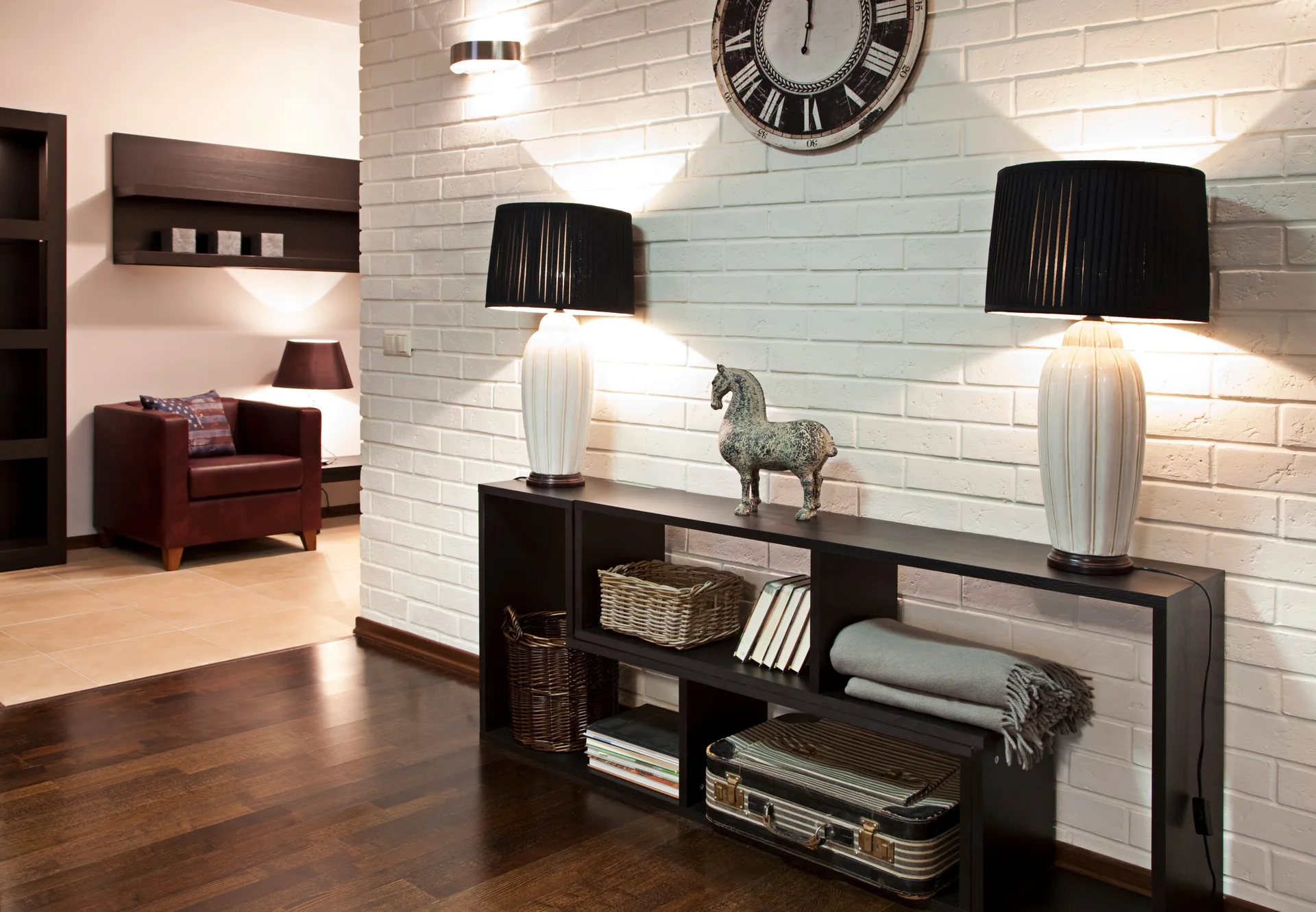 A dark wood console table with symmetrical black-shaded lamps and a horse sculpture stands against a textured white painted brick wall.