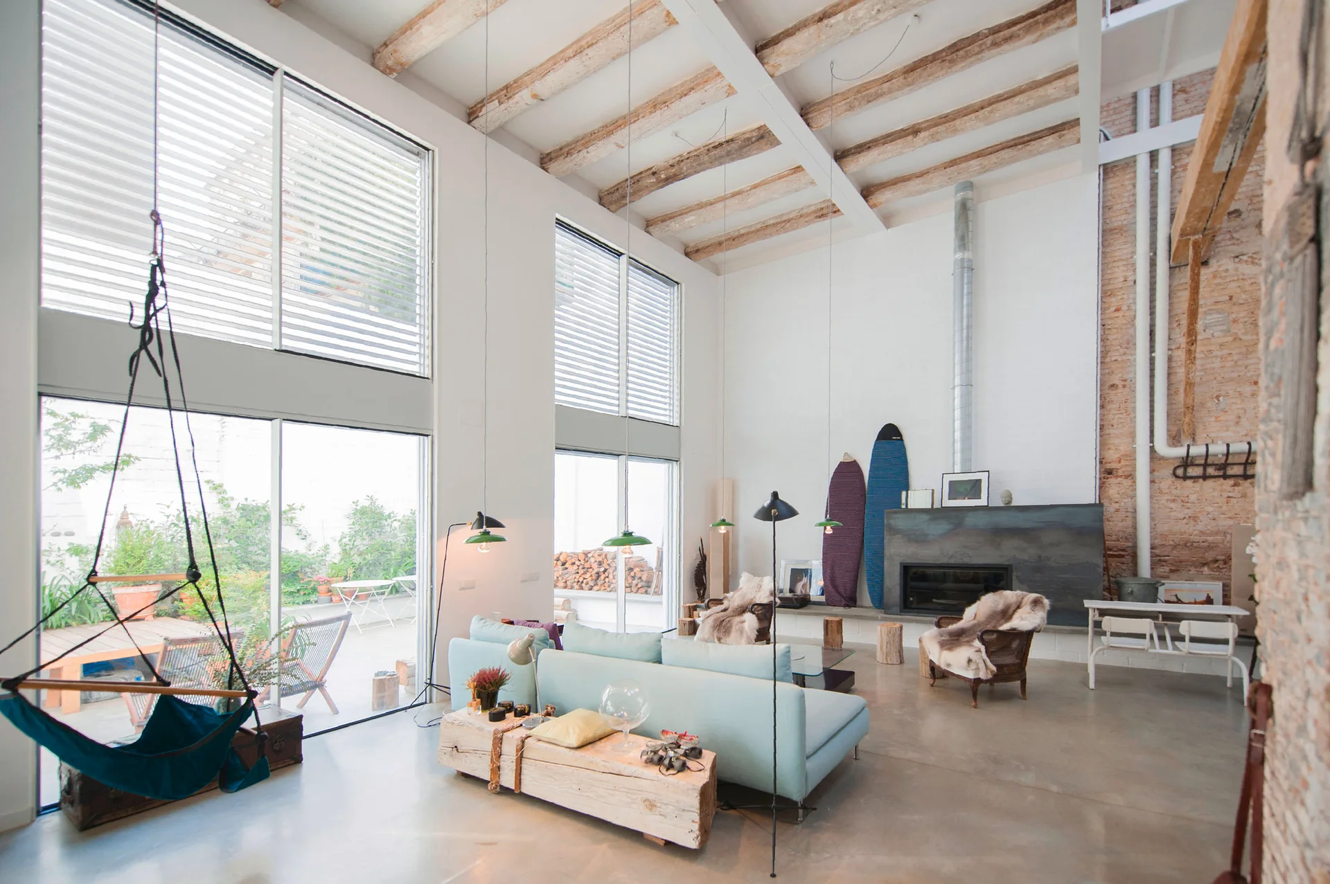 A spacious loft living room featuring a light blue sofa, exposed wooden ceiling beams, large windows, and surfboards resting against a white wall.