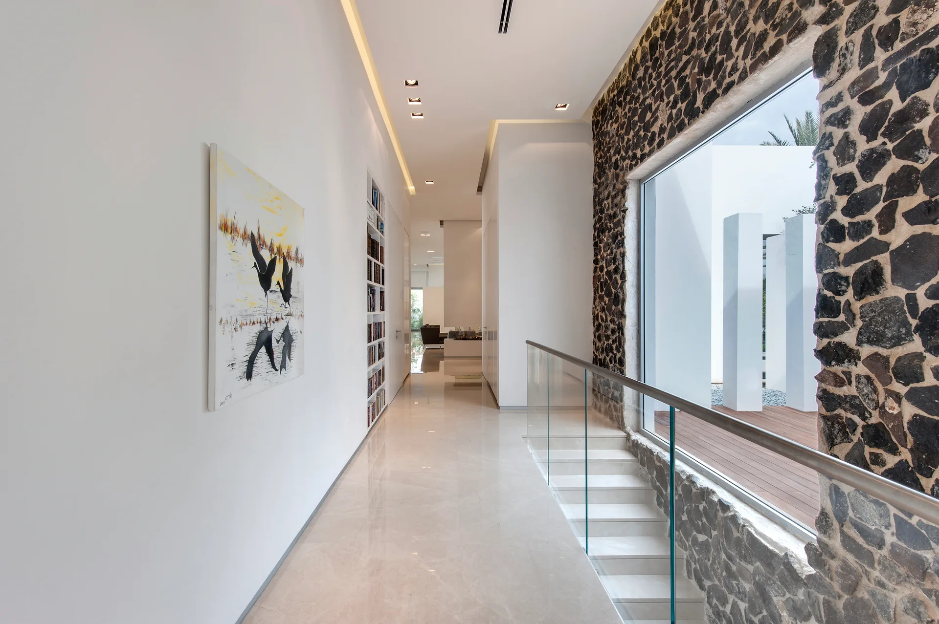 A modern hallway features polished beige tile floors, white walls with a recessed bookshelf, and a staircase with a glass railing against a stone wall.