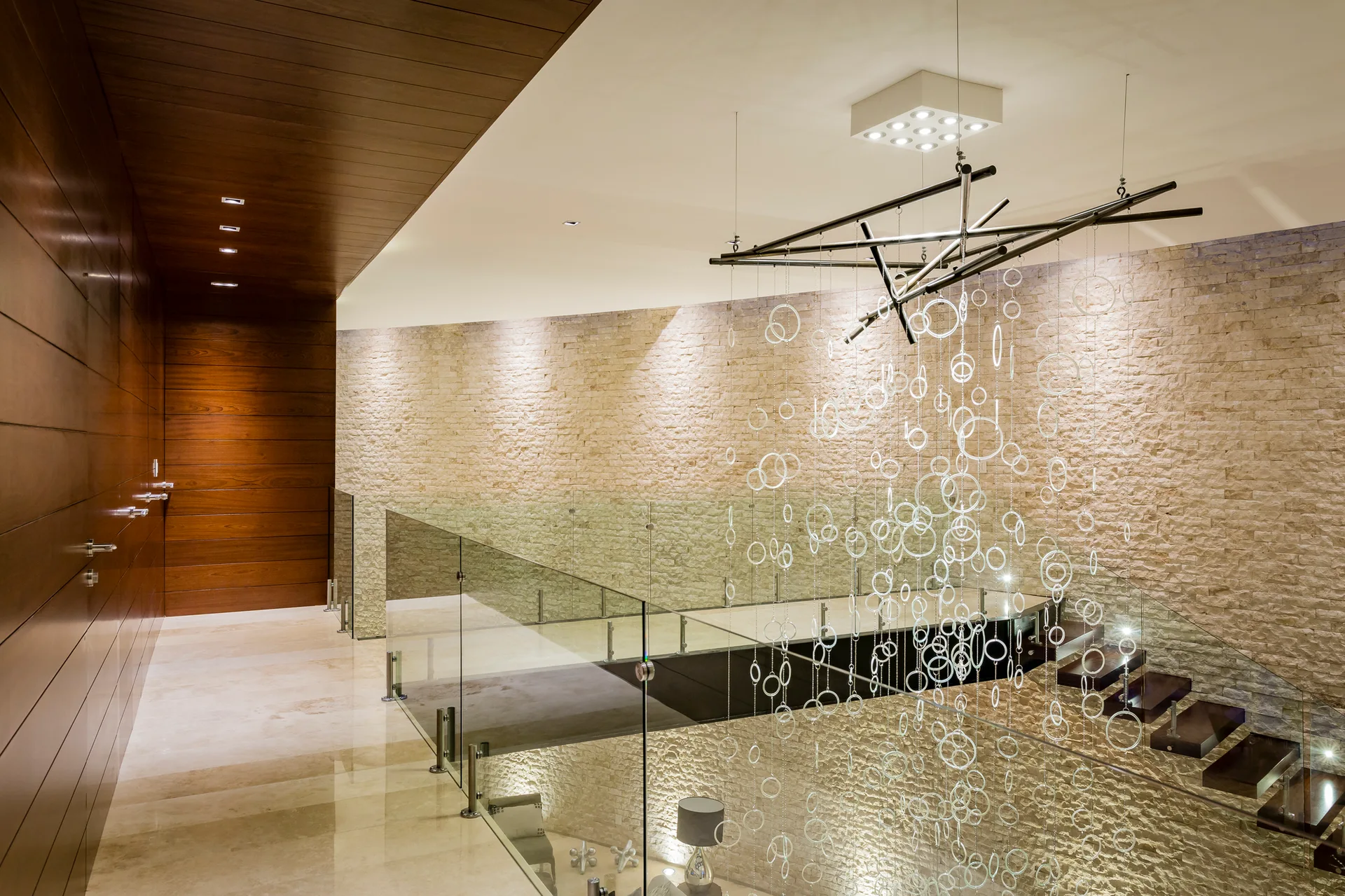 A modern staircase features floating dark wood treads, a glass railing, a textured stacked stone wall, and a cascading ring chandelier.