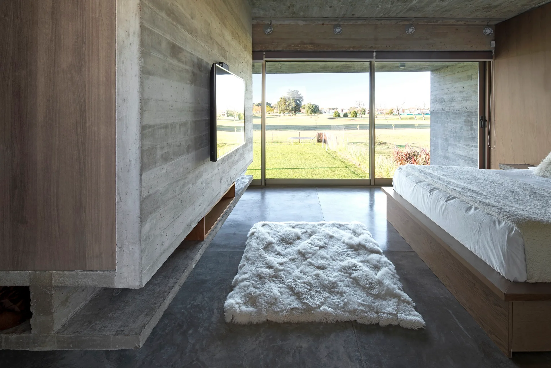 A minimalist bedroom features board-formed concrete walls, a low wood platform bed, a shaggy white rug, and sliding glass doors opening to a grassy field.