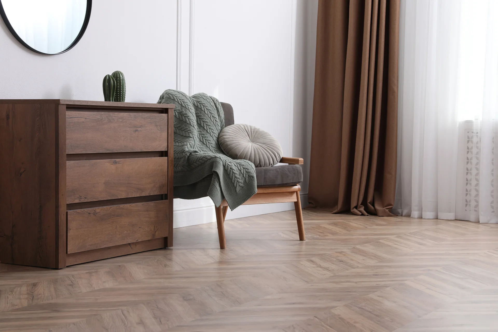 A wooden dresser and an armchair with a green knit throw sit on a herringbone wood floor next to brown curtains.