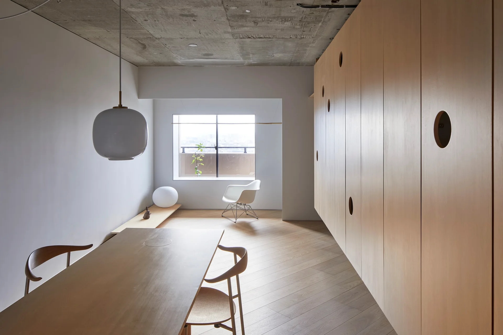 A minimalist dining room with a raw concrete ceiling, herringbone wood floors, a wooden dining table, and built-in wood cabinets with circular cutouts.