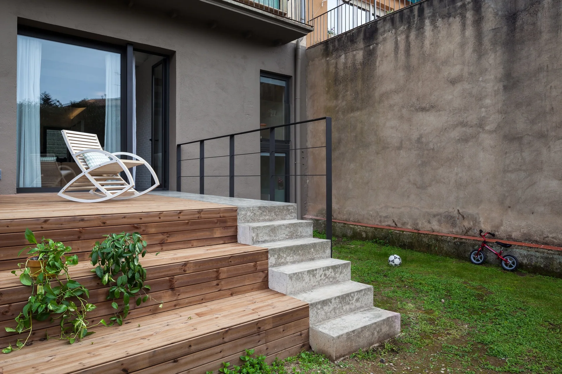 A multi-level wooden deck with a white rocking chair features cast concrete steps leading down to a grassy yard next to a stucco exterior wall.