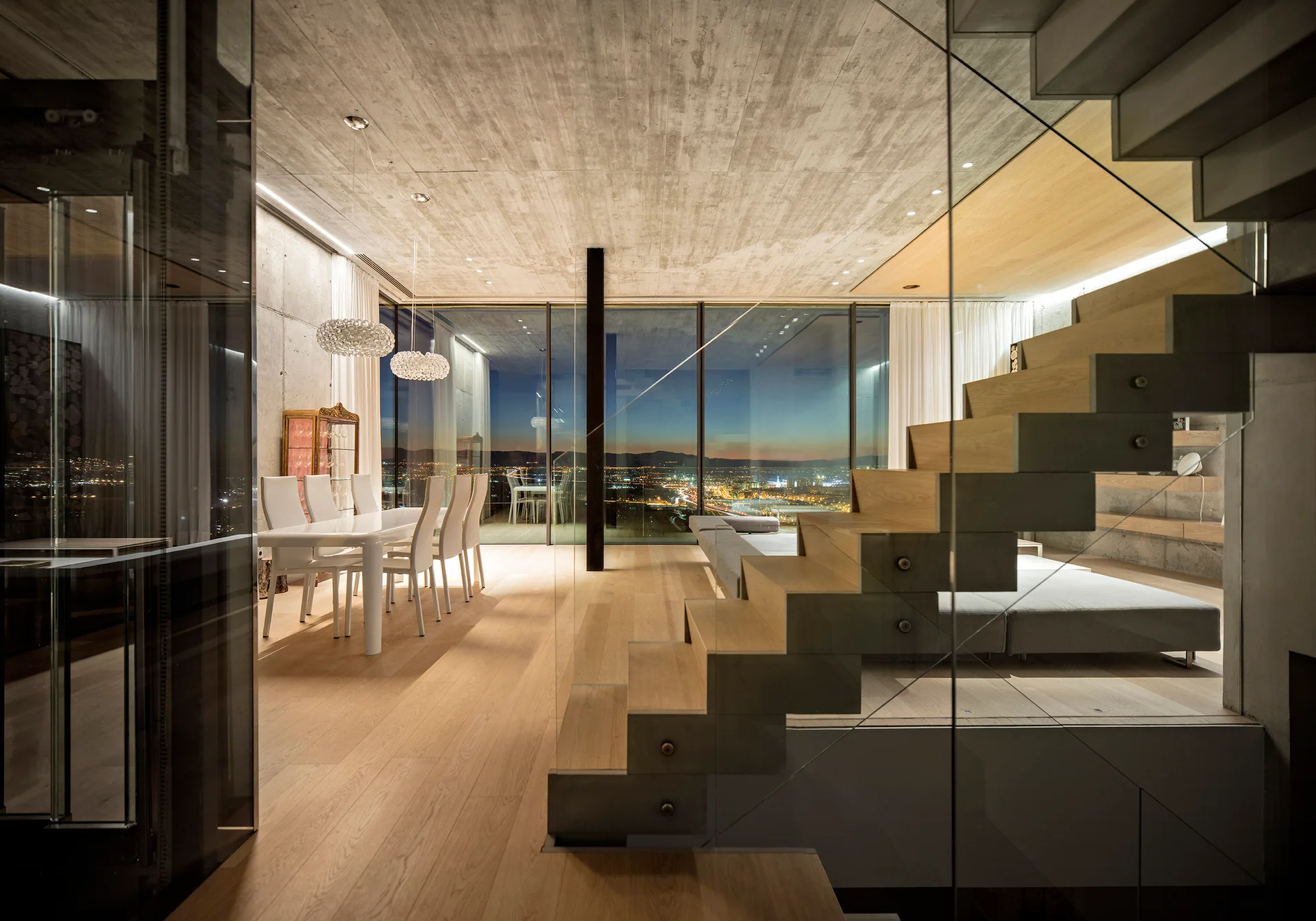 A modern open-plan living and dining space featuring a wood staircase behind glass, concrete ceilings, and floor-to-ceiling windows showing a night skyline.