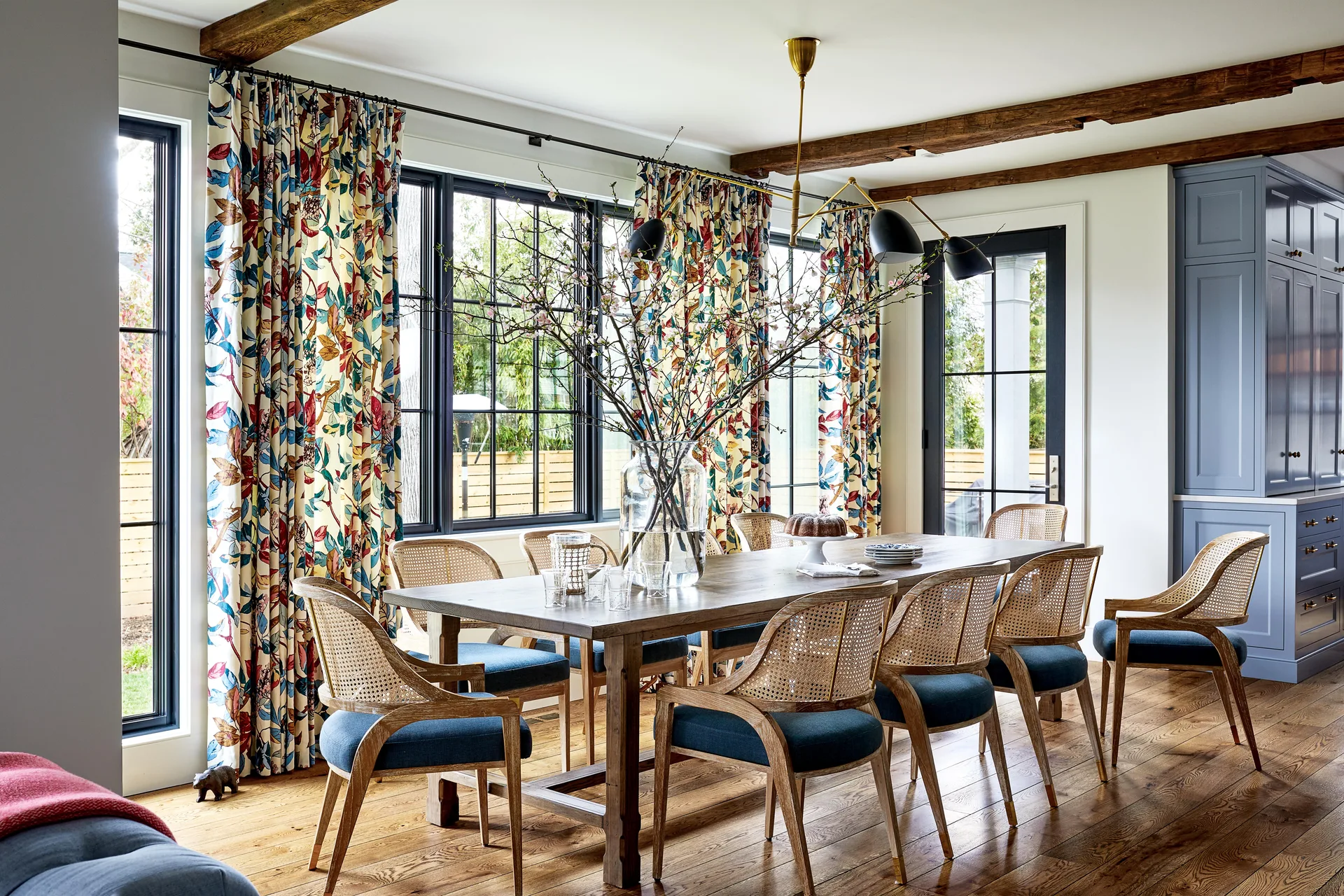 A dining room featuring a wood table, cane chairs with blue cushions, floral curtains, and dark ceiling beams.