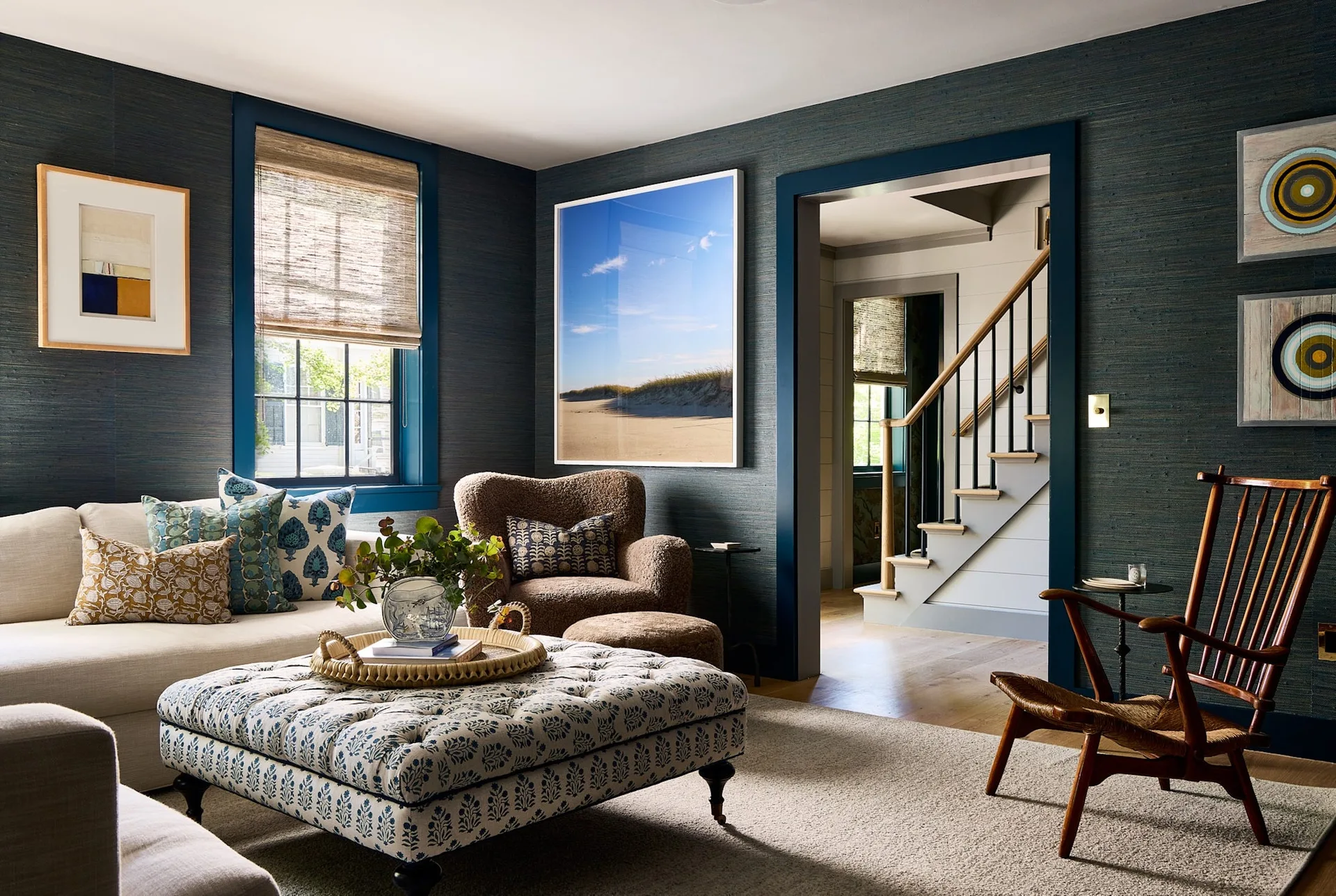 A cozy living room featuring deep blue grasscloth walls, a cream sofa, a large tufted patterned ottoman, and a brown armchair.