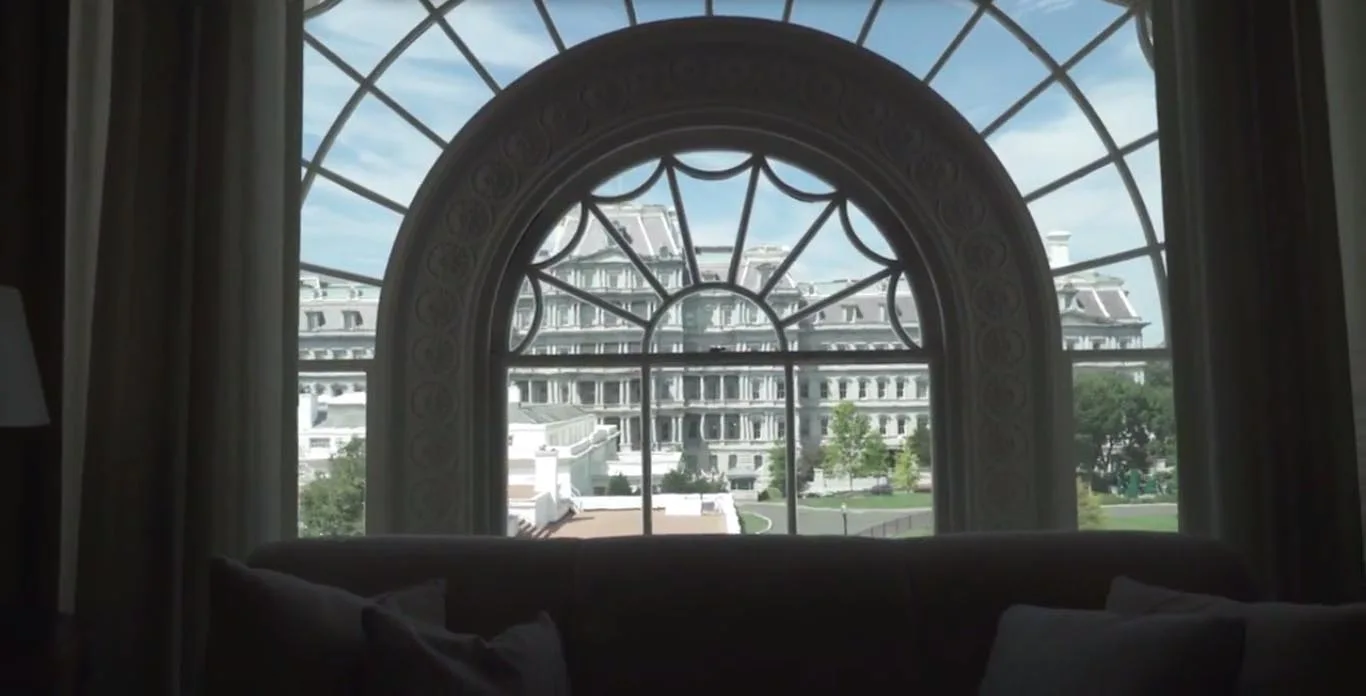 A dark sofa with throw pillows sits in front of a large arched window with detailed mullions overlooking a classical stone building.