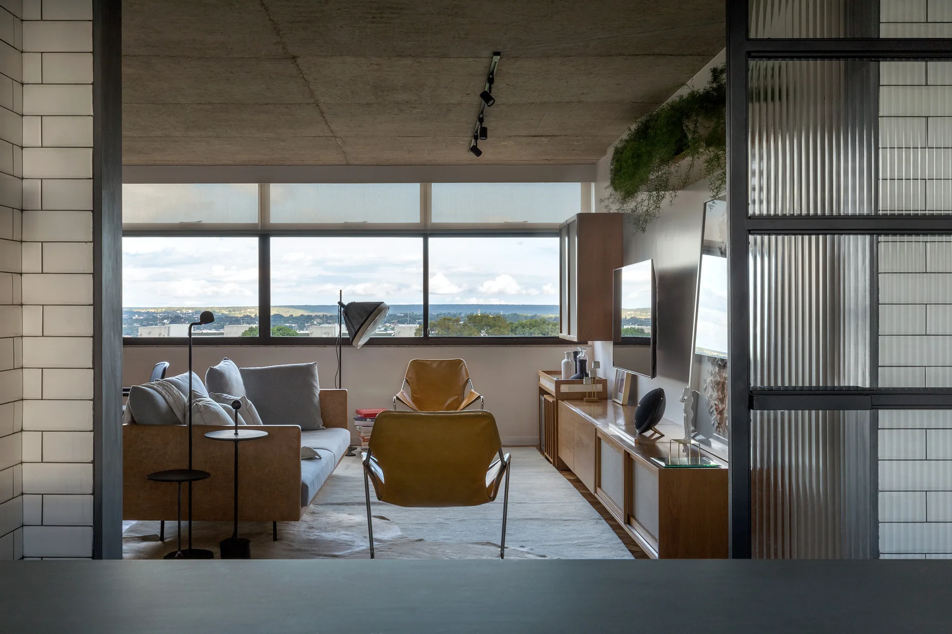 An industrial living room features tan leather armchairs, a grey sofa, a wooden media console, and an exposed concrete ceiling under track lights.