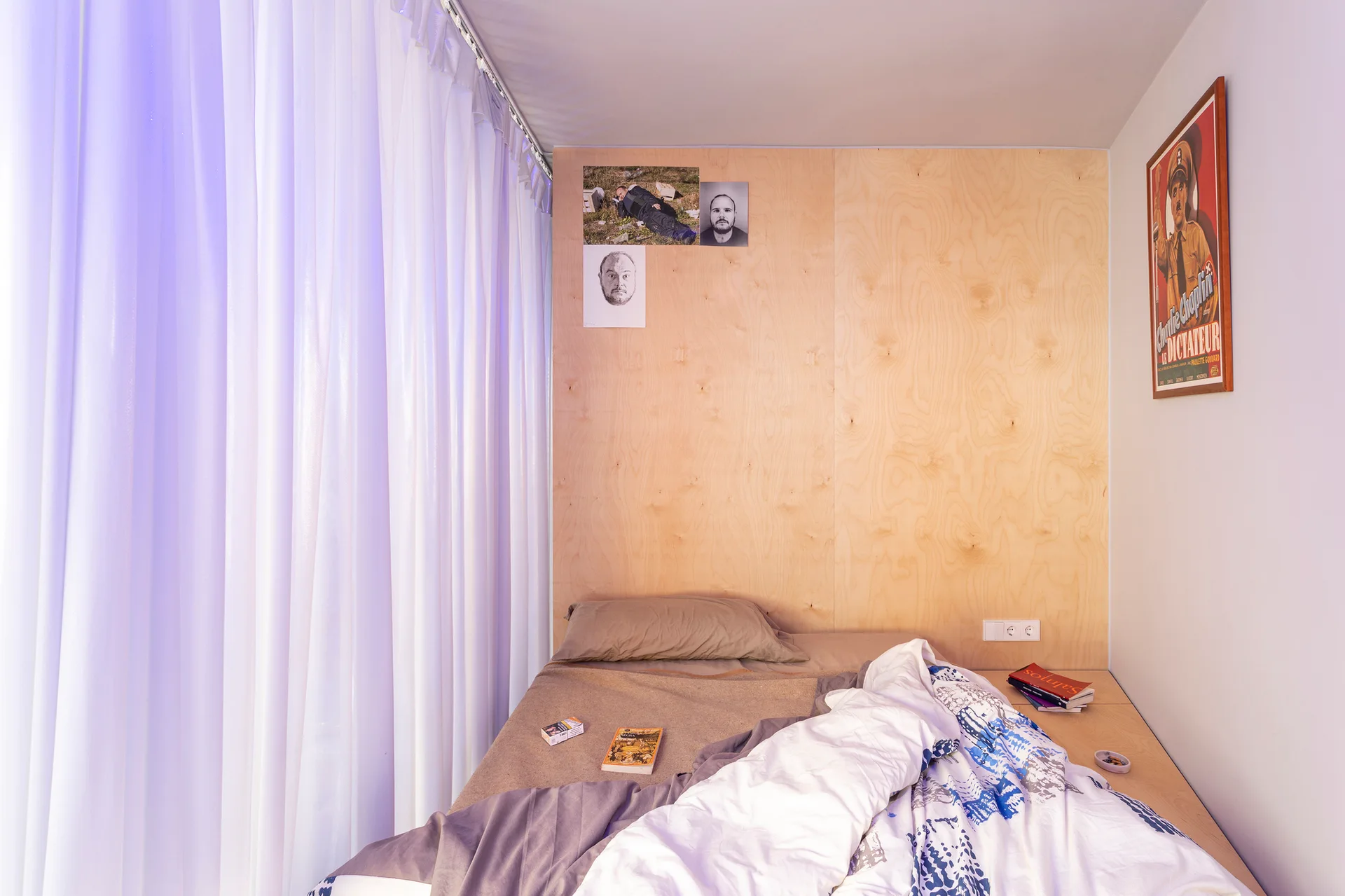 A platform bed with beige and blue linens is tucked against a light-colored plywood paneled wall beside sheer white curtains.
