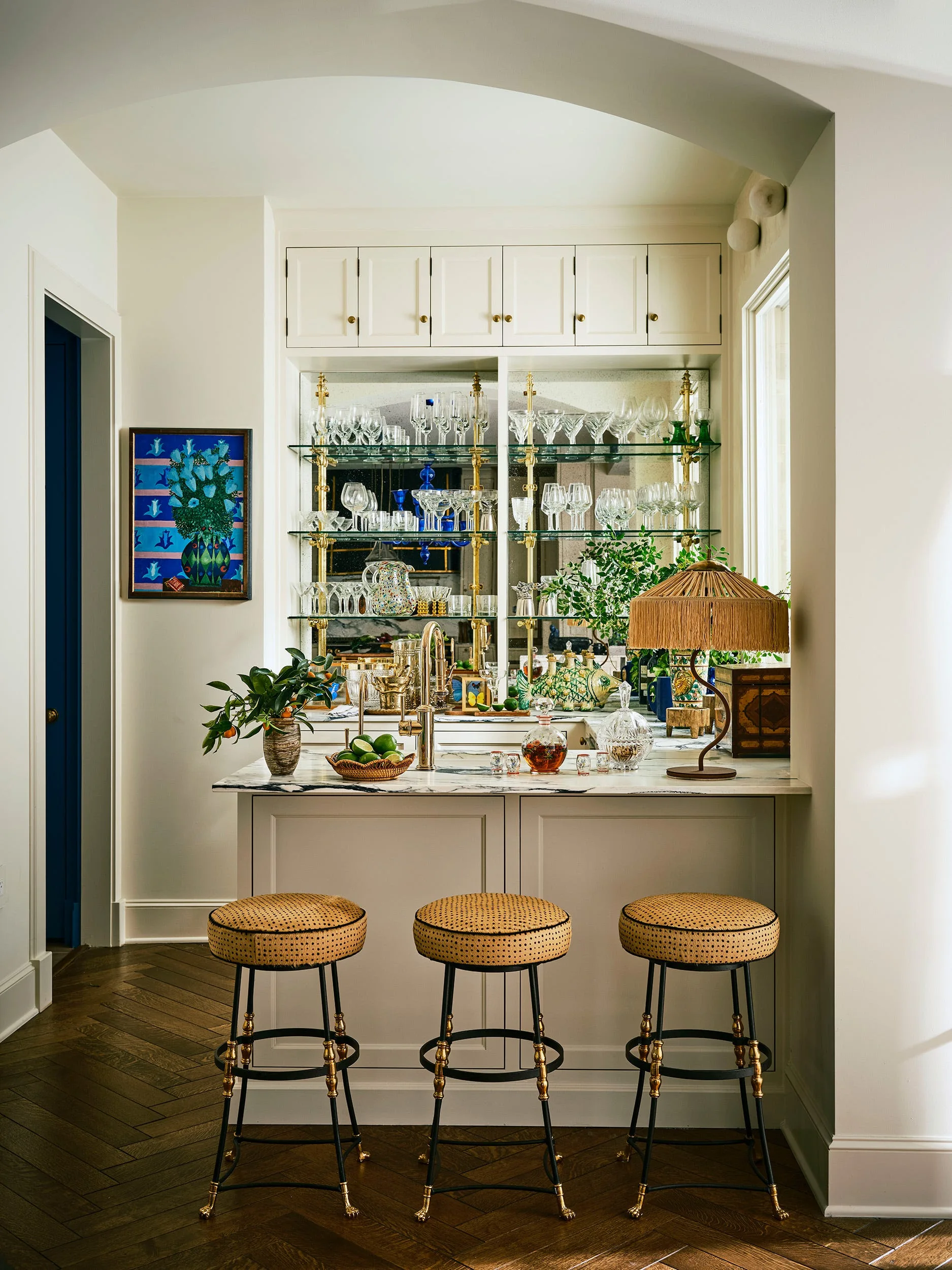 A built-in home bar features a marble-topped counter, three rattan-seated stools, and a glass-shelved display with glassware against a mirrored back.