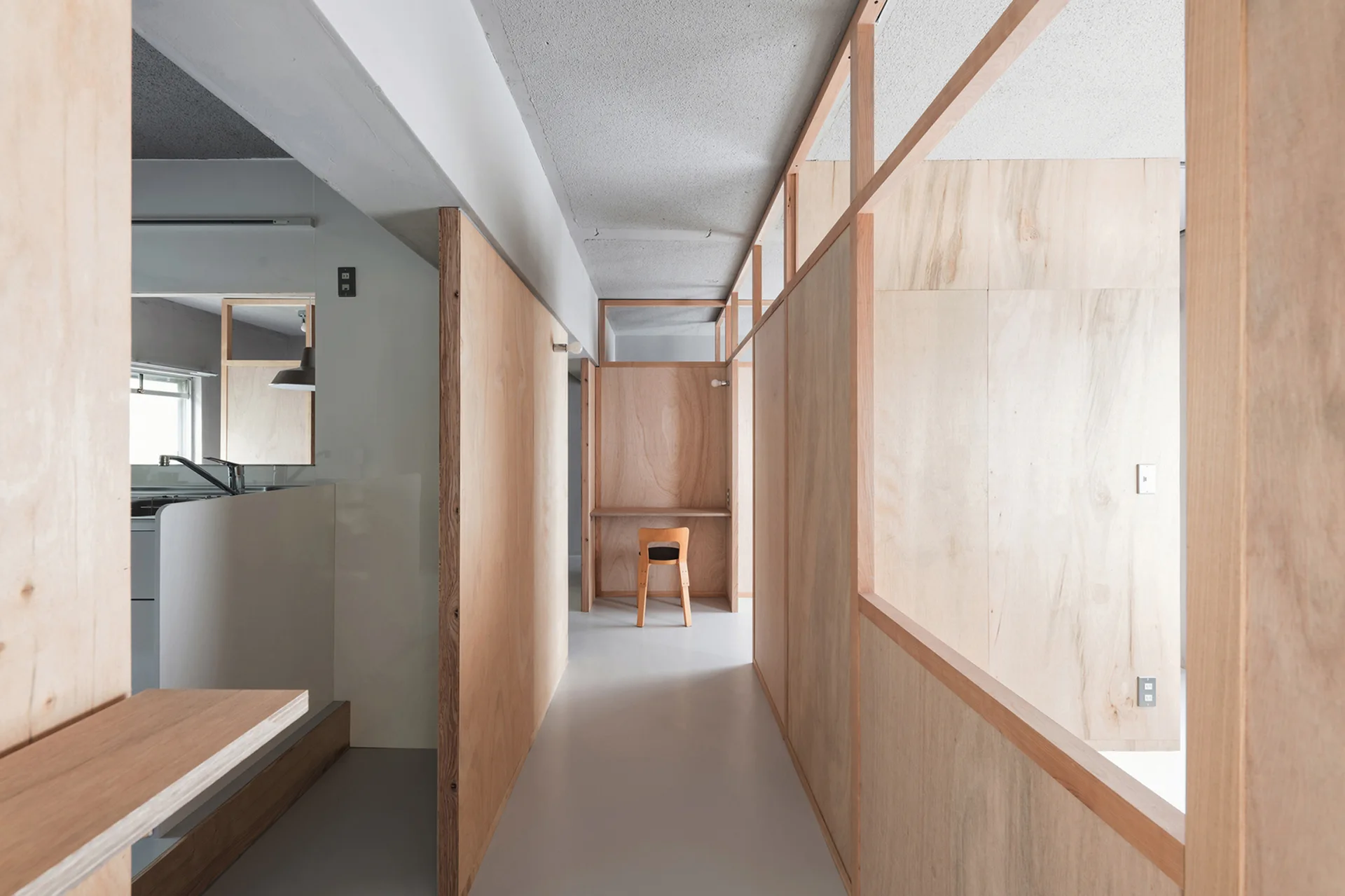 A bright hallway features plywood partition walls, an exposed concrete ceiling, and a built-in desk with a wooden chair at the far end.