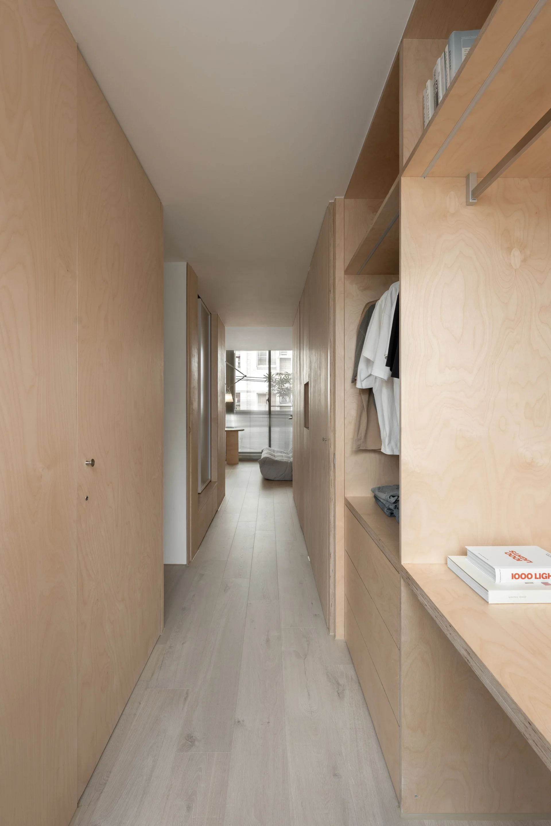 A minimalist dressing corridor featuring light-colored plywood built-in wardrobes and open shelving leading to a sunlit room in the distance.