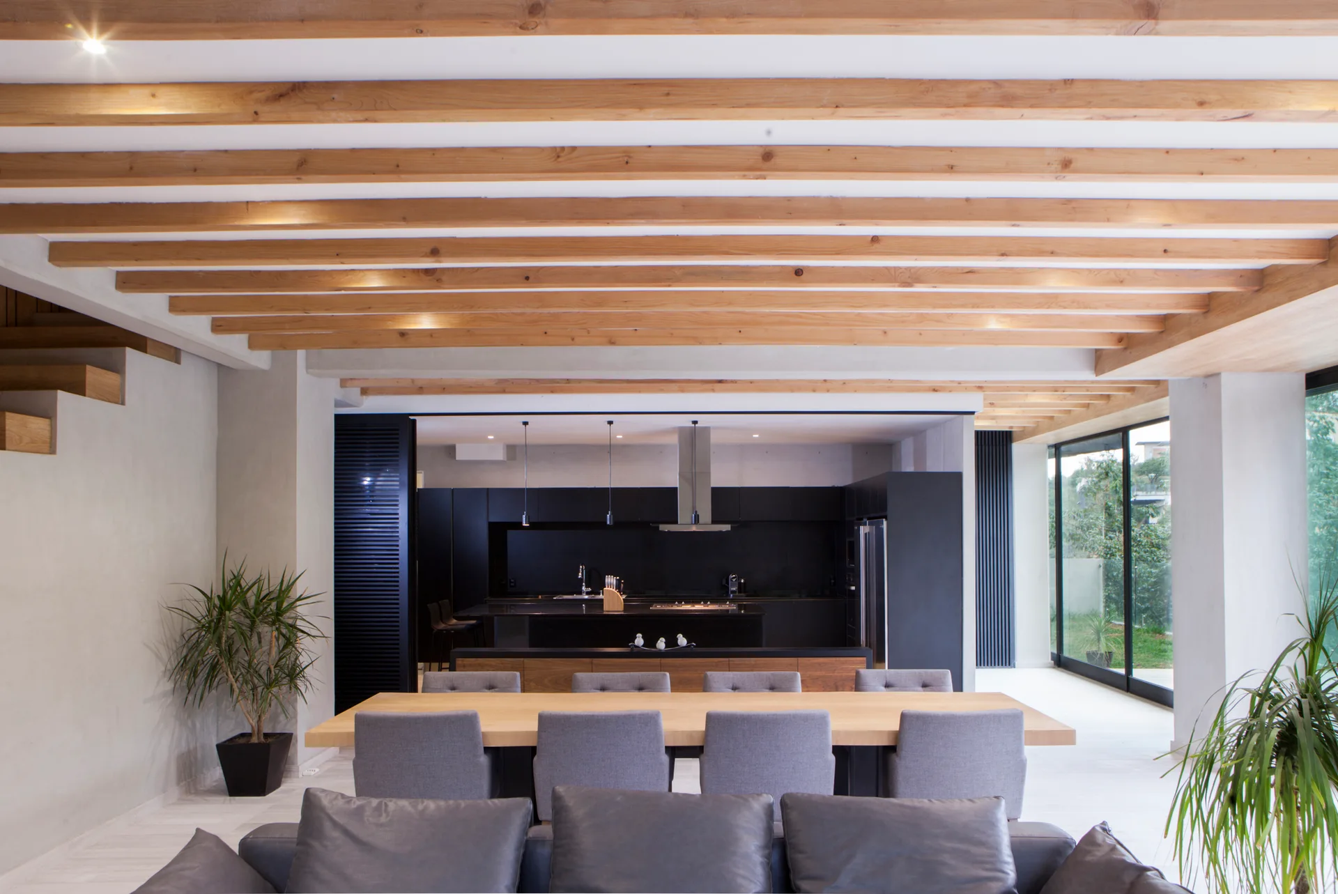 An open-plan dining space features a long wooden table with grey chairs beneath a ceiling defined by repetitive exposed wood beams.