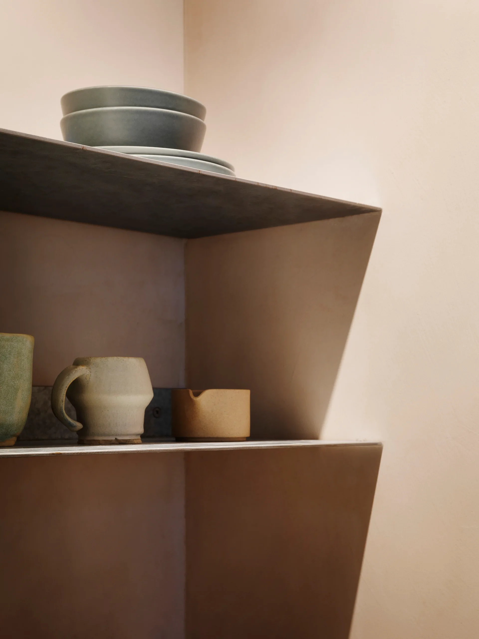Handcrafted ceramic mugs and bowls sit on minimalist metal shelves mounted in a matte beige wall corner.