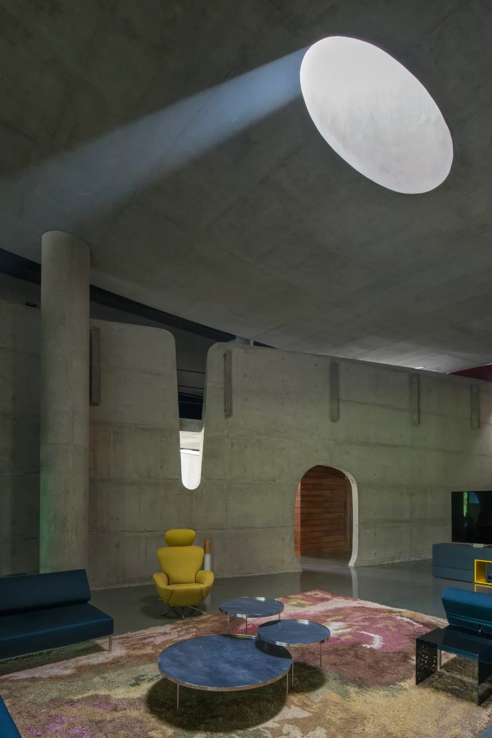 A minimalist living room featuring raw concrete walls, a large circular skylight, a patterned pink rug, and a yellow sculptural armchair.
