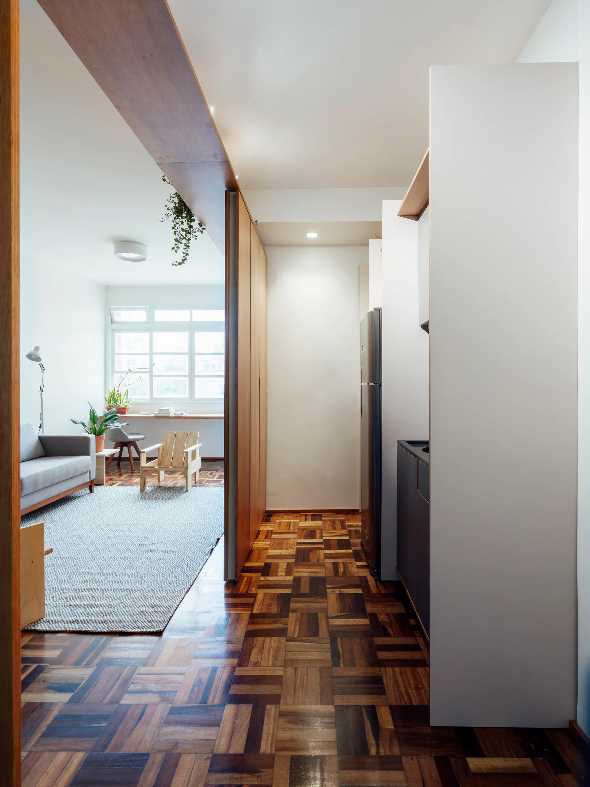 A bright living room with a grey sofa and parquet flooring leads into a compact kitchen area framed by a wood beam ceiling header.