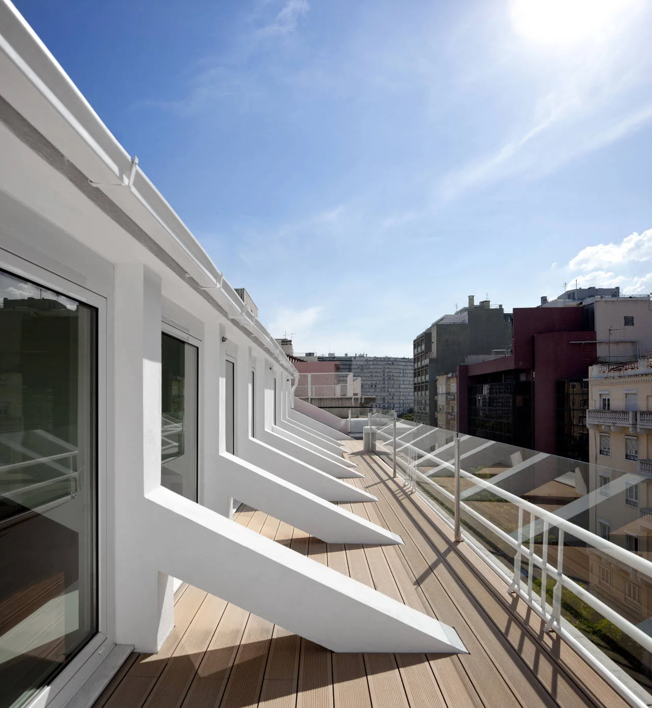 A long outdoor balcony features light wood-toned decking planks, a transparent glass railing, and a row of white angled structural supports.