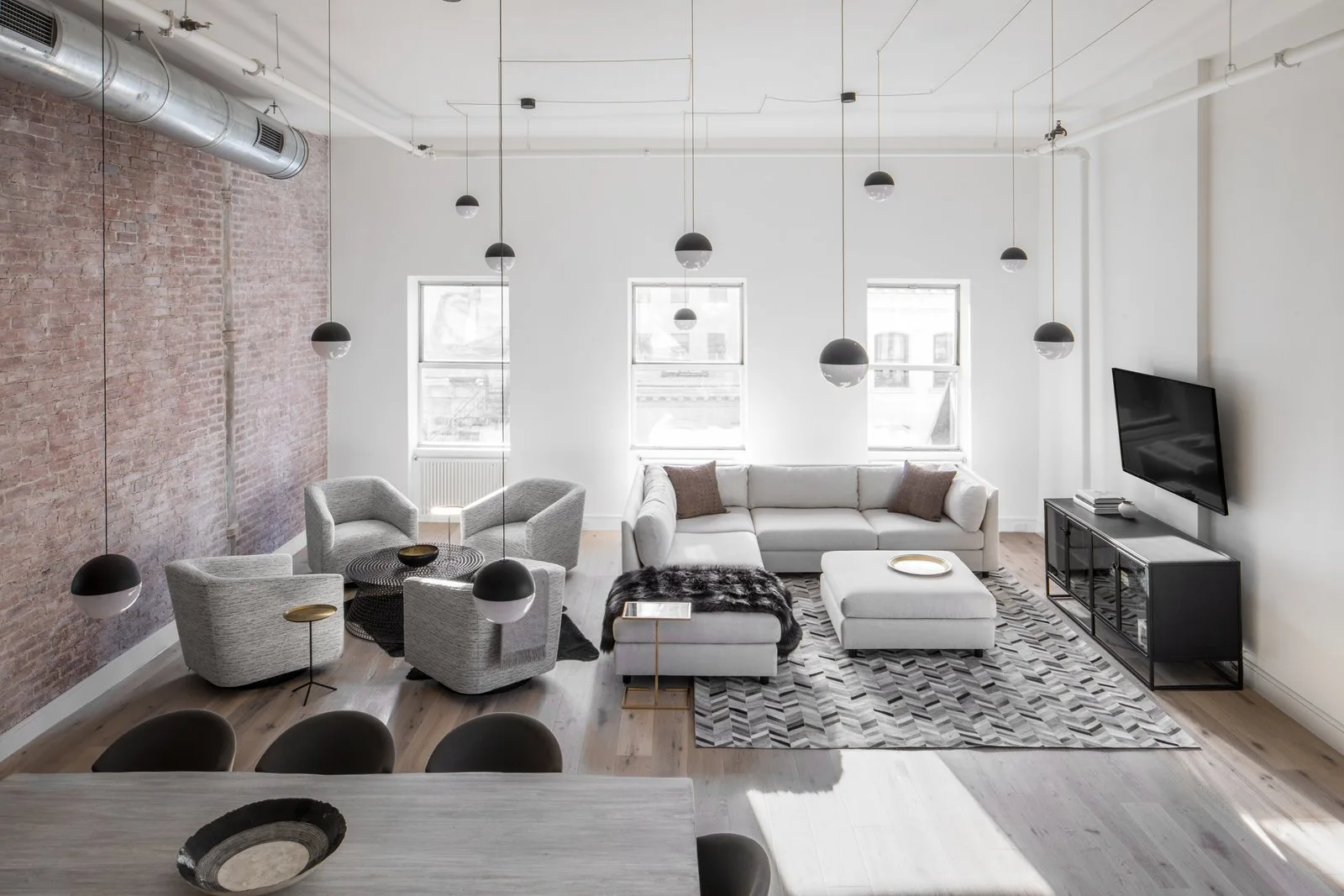 An open-plan loft living room featuring a light grey sectional sofa, four armchairs, an exposed brick wall, and multiple hanging globe pendant lights.