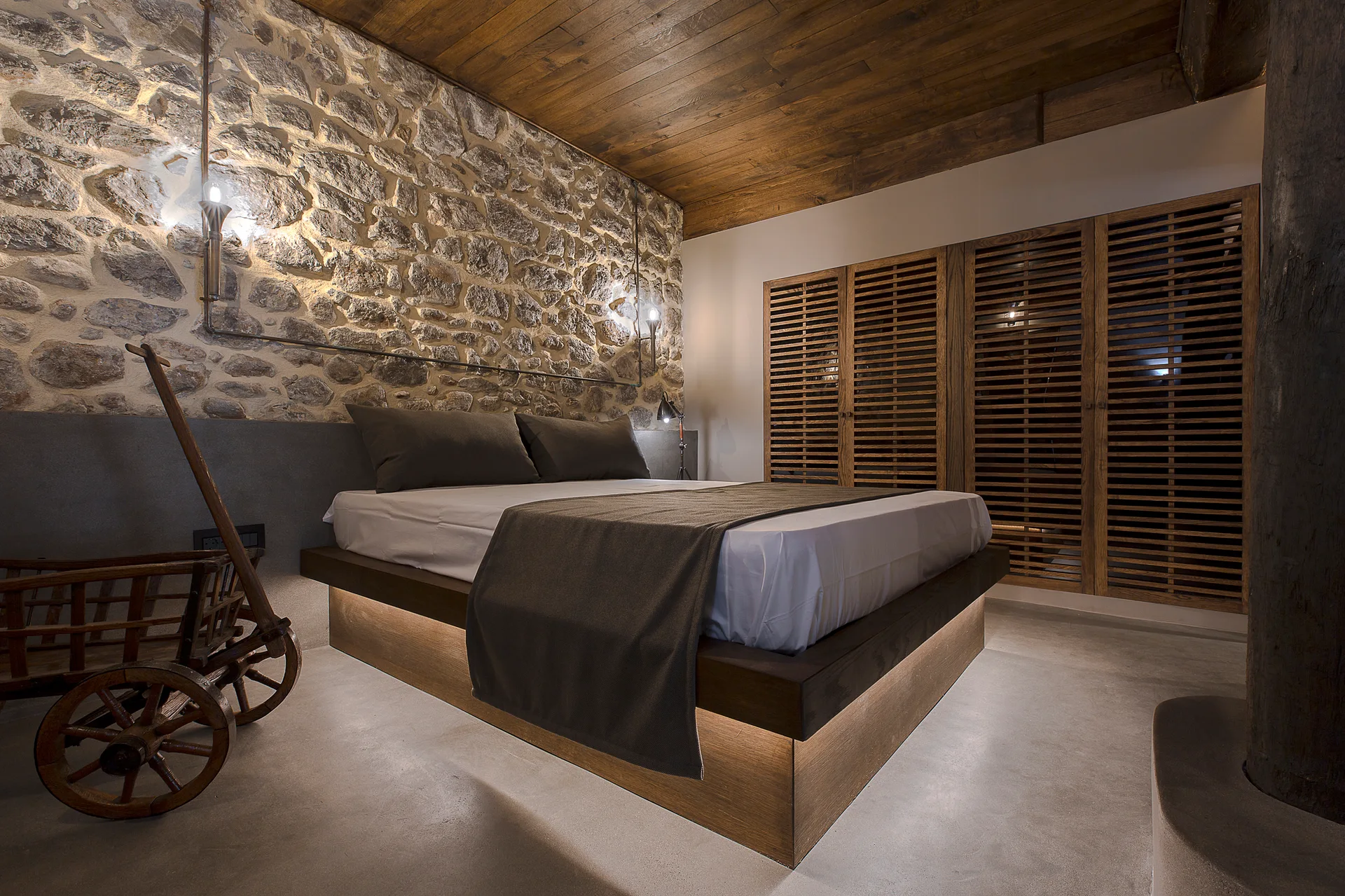A rustic bedroom features a platform bed with under-bed lighting positioned against a textured stone wall next to slatted wood closet doors.
