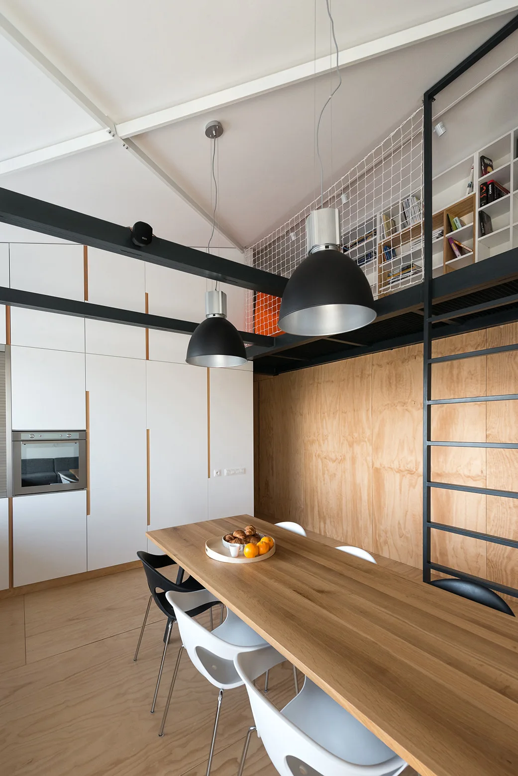 A dining room features a wood table with black and white chairs beneath a mezzanine loft with black steel beams and hanging dome lights.