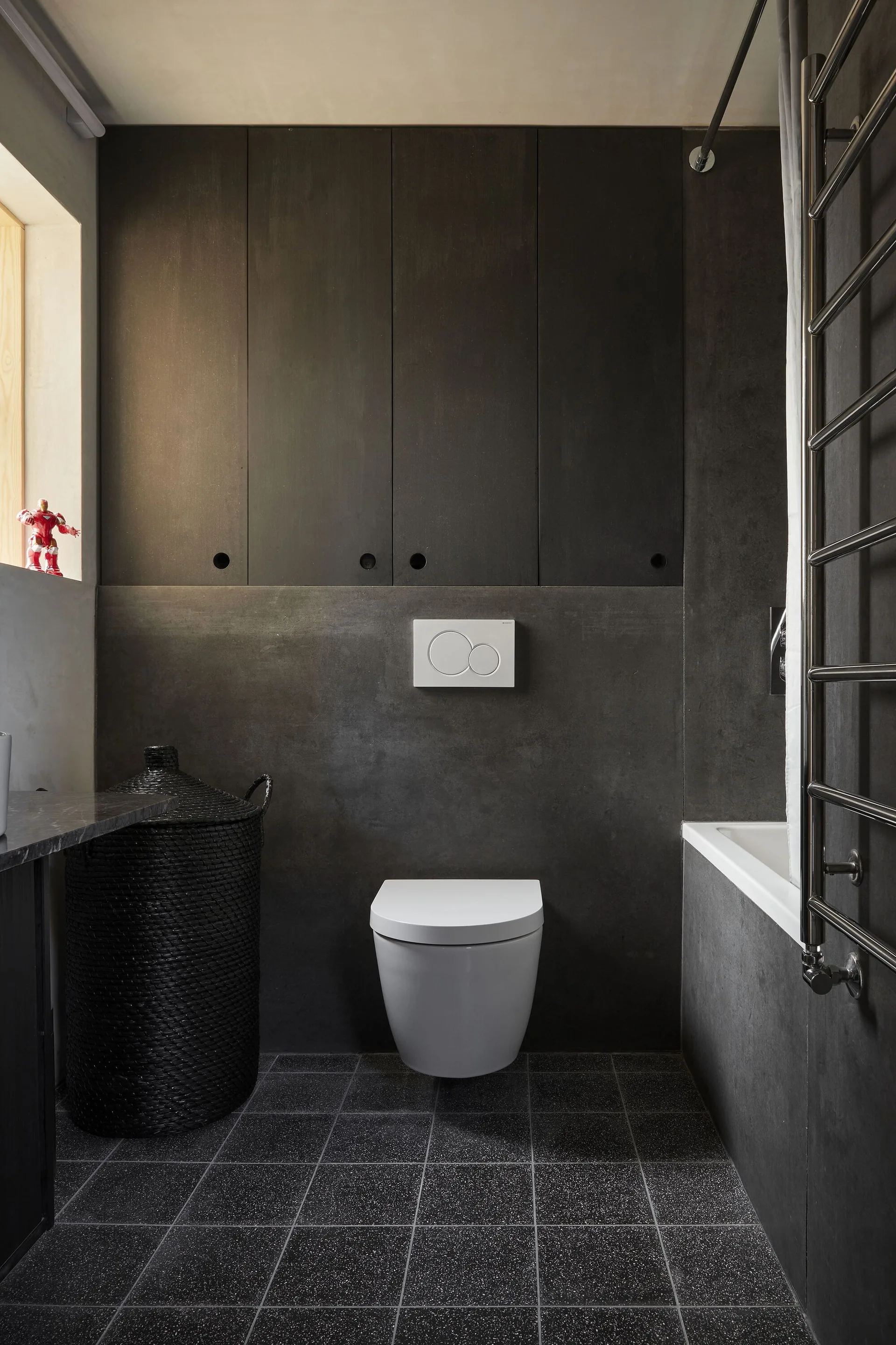 A contemporary bathroom features dark charcoal wall panels, a white wall-mounted toilet, dark tiled floors, and a black wicker laundry hamper.