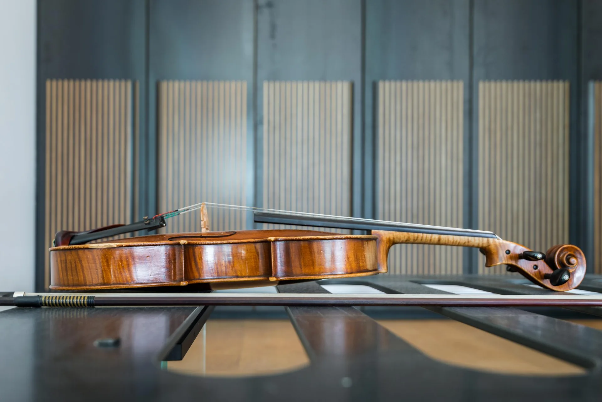 A polished wooden violin and its bow lie horizontally on a dark slotted bench in front of a wall with vertical wood slats.