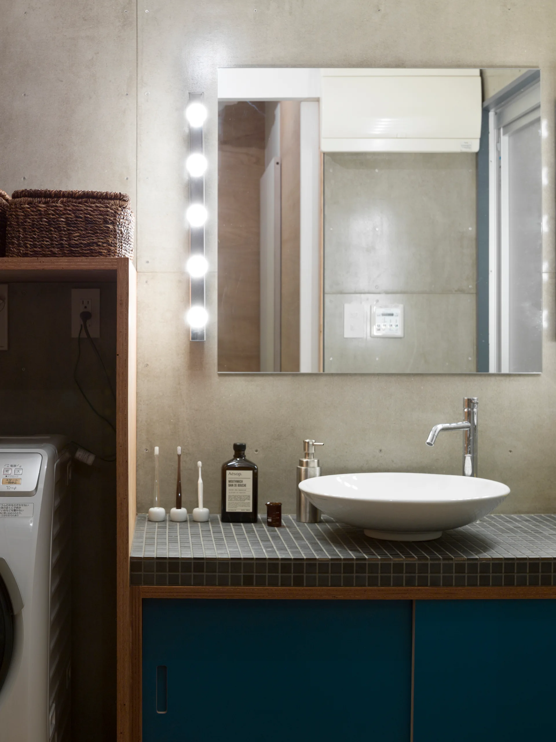 A modern bathroom vanity features a white vessel sink on a dark mosaic-tiled countertop with teal sliding cabinets set against a concrete wall.